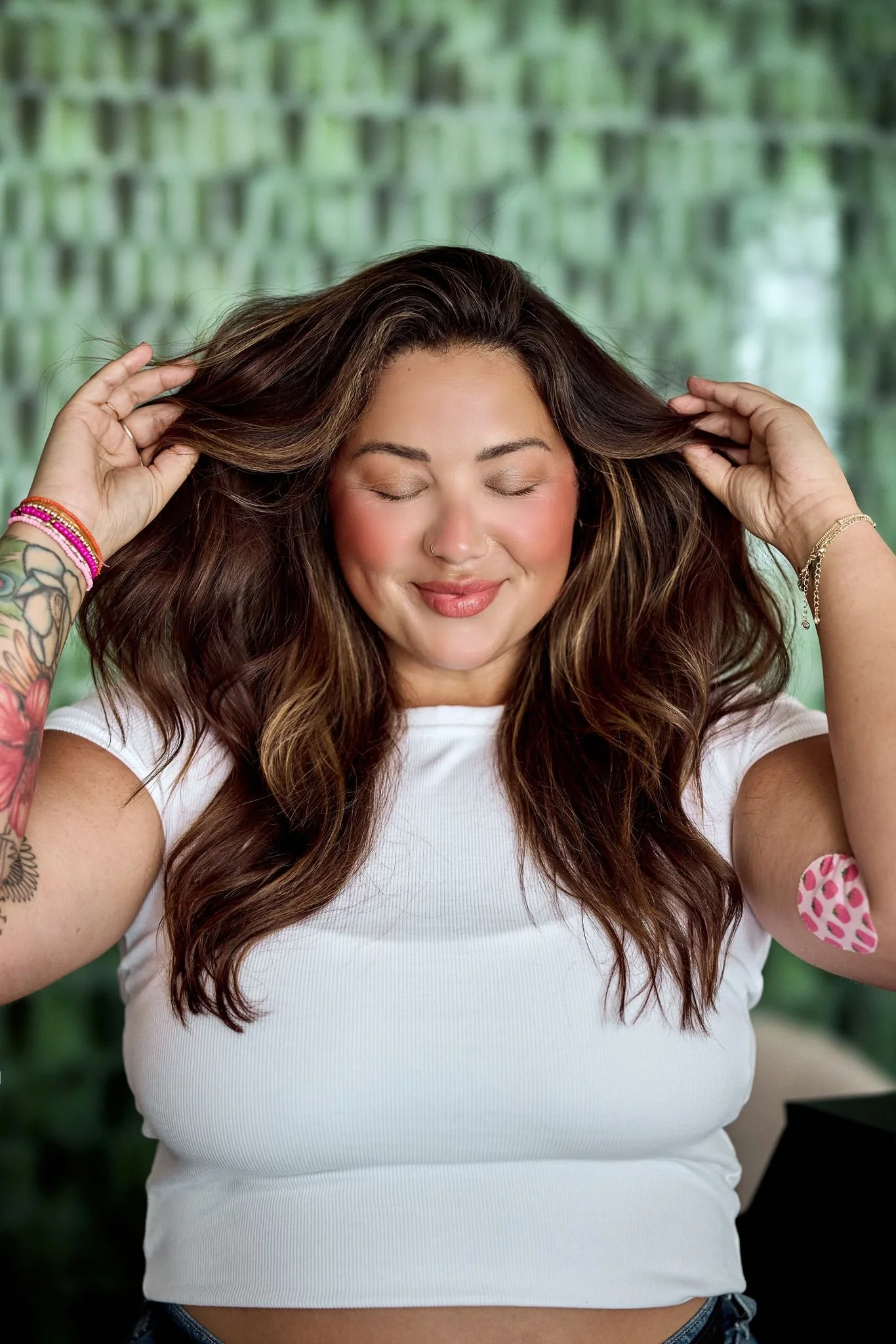 A woman with long wavy brown hair and light skin is smiling with closed eyes. She is holding her hair with both hands, which are adorned with colorful bracelets. She is wearing a white t-shirt, and there are tattoos on her left arm. The background appears to be a green outdoor setting with a wooden fence.
