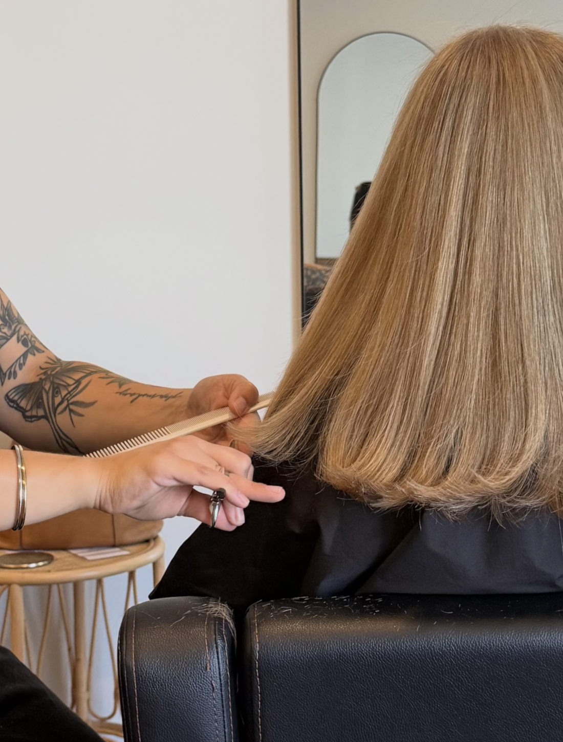 A woman with long, blonde hair is getting her hair styled by a stylist, who is using a comb. The stylist has tattoos on her arm. The scene takes place in a salon with a mirror visible in the background.