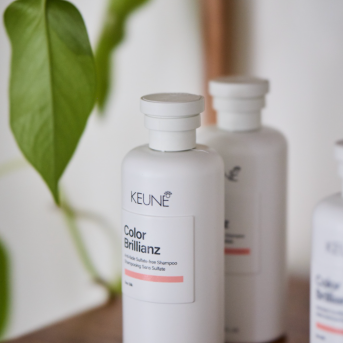Two white bottles of KEUNE Color Brillianz shampoo on a wooden surface, with green leaves in the background.