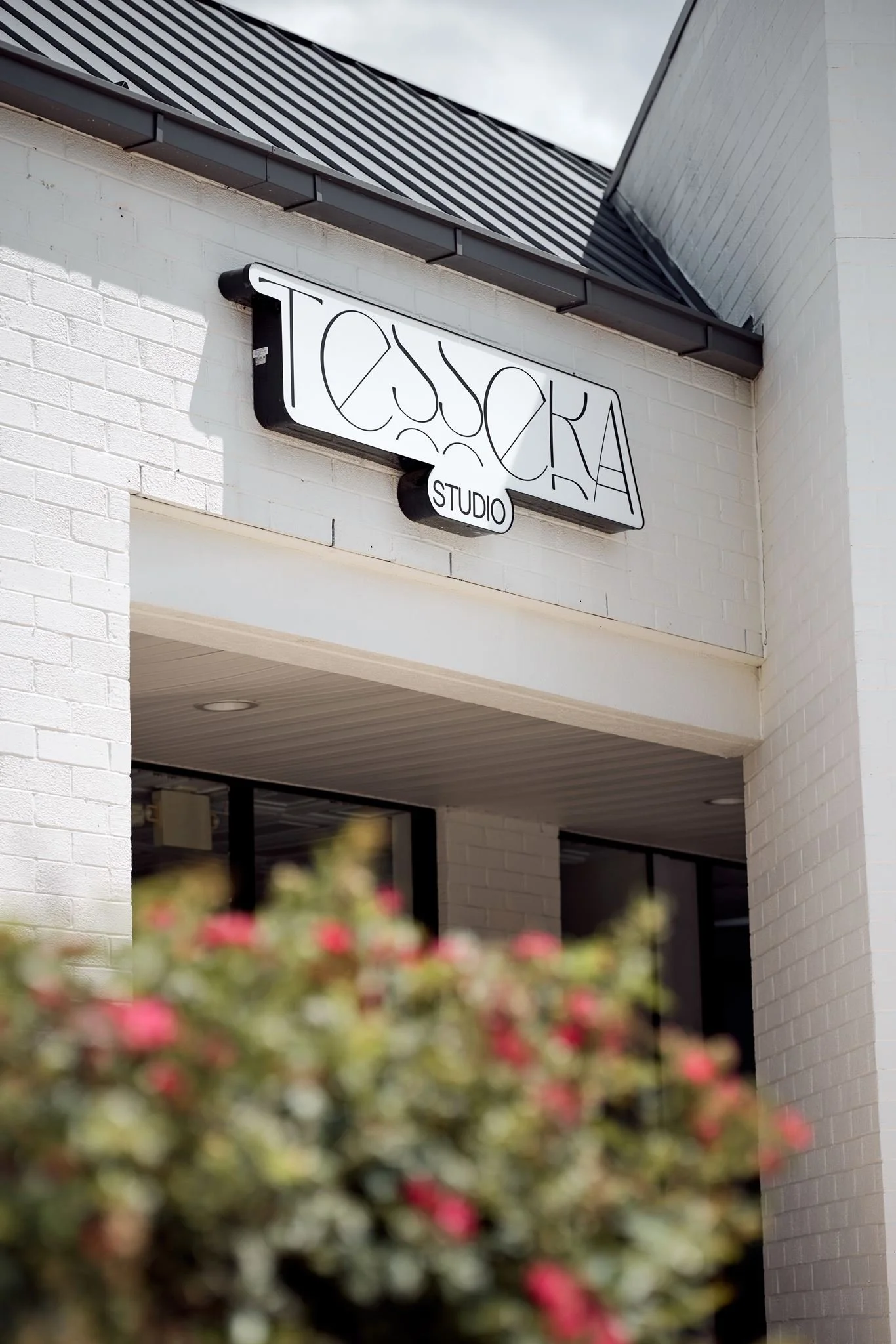 Exterior of a building with a sign that reads 'TOSKA STUDIO'. The sign is mounted on a white brick wall beneath a gray metal roof with black trim. Blurred pink flowers are visible in the foreground.
