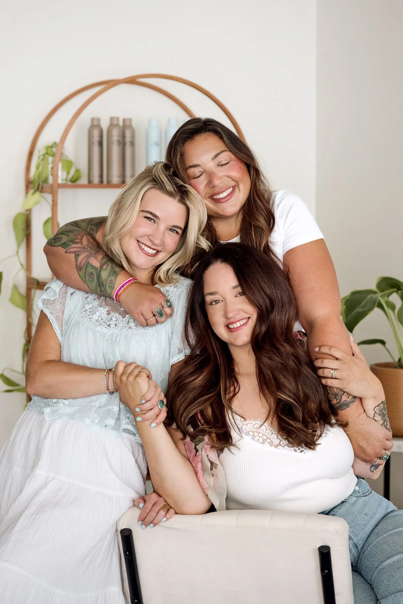 Four women with various hair colors and styles, smiling and hugging, sitting together in a bright room with houseplants and a shelf with bottles in the background.