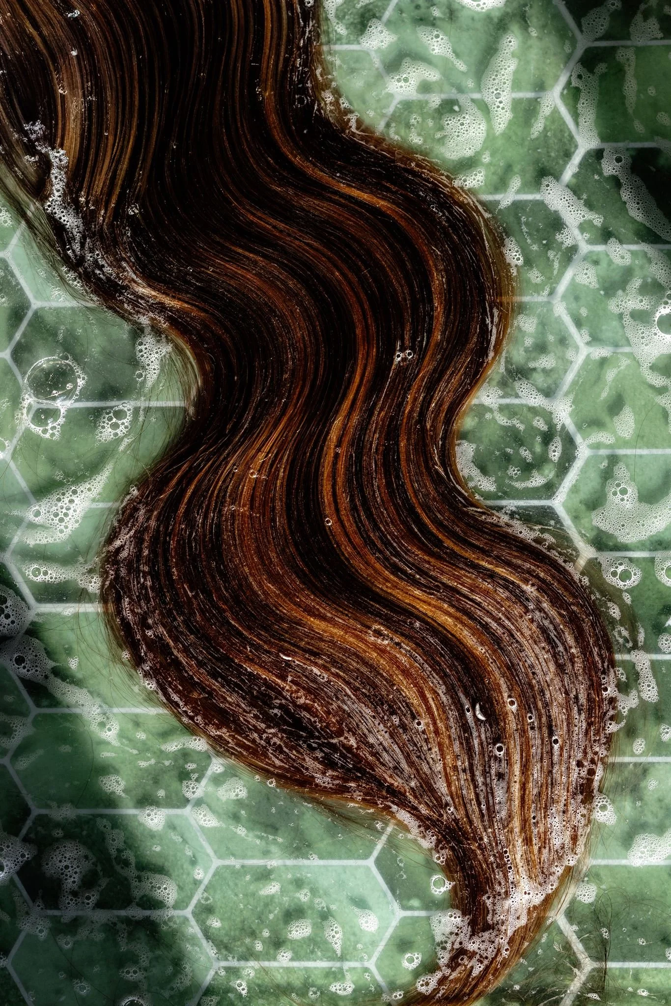Close-up of a brown hair strand in a bath with soap bubbles on a green hexagonal patterned surface.