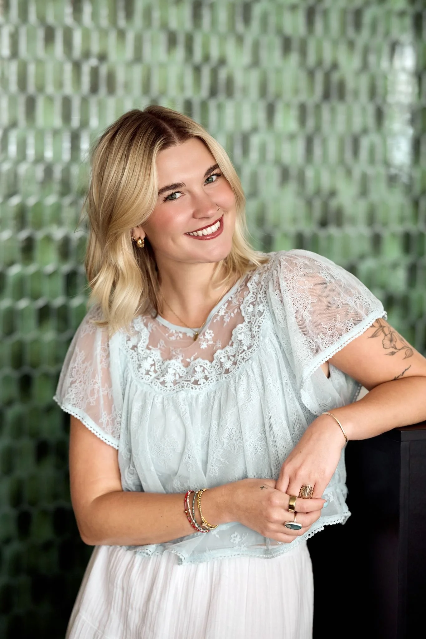 A woman with blonde hair styled in waves, smiling and wearing a white lace blouse with jewelry, standing in front of a green geometric patterned background.