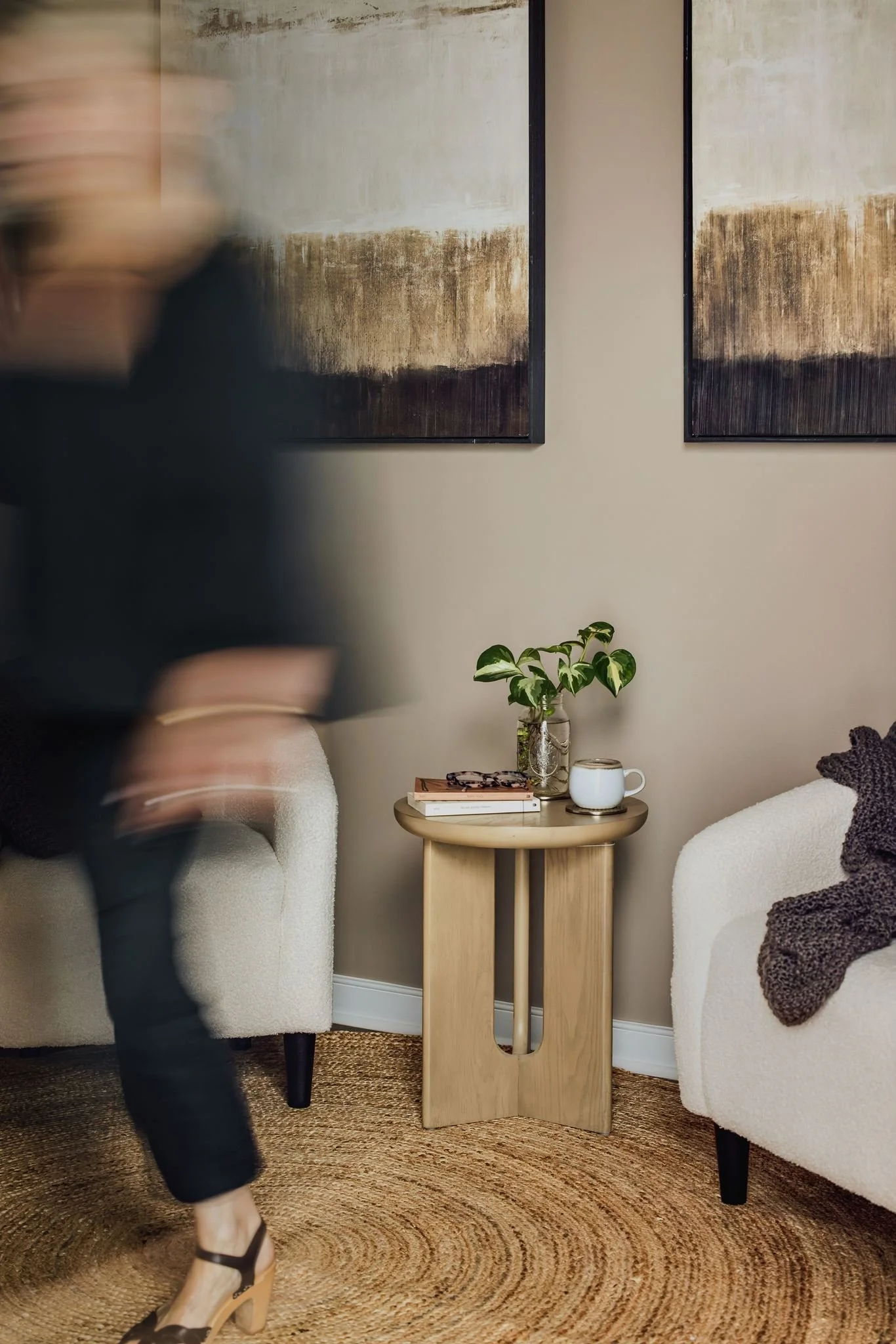 A cozy living room with two white armchairs, a small round wooden table with books, a plant, a coffee mug, and a textured area rug. Two abstract paintings hang on the wall.