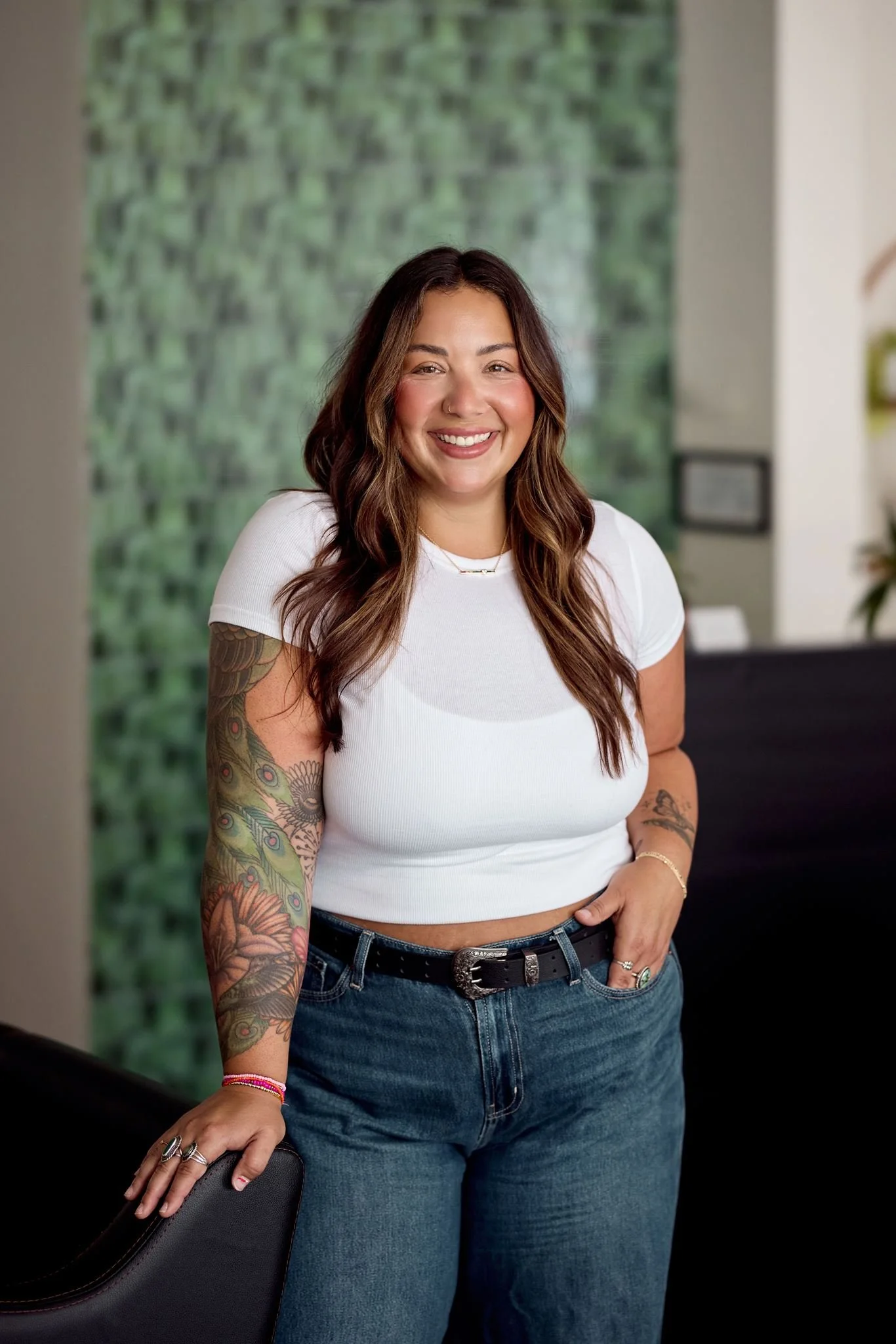 A woman with long brown hair, smiling, wearing a white crop top, blue jeans with a black belt, and tattoos on her left arm. She is standing indoors, holding a black chair with her right hand, with a green leafy background.