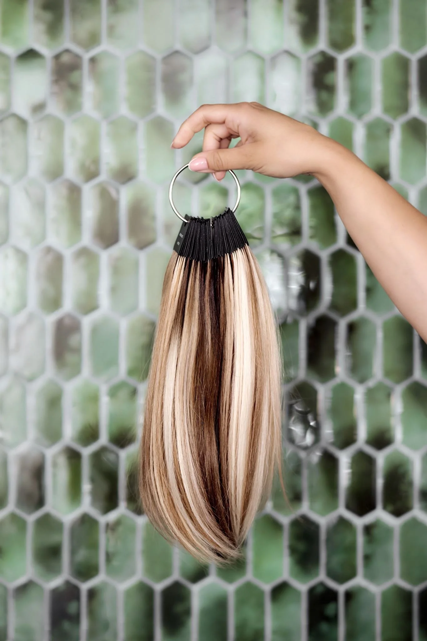 Hand holding a circular hair color ring with blonde hair samples against a hexagonal wire mesh background.