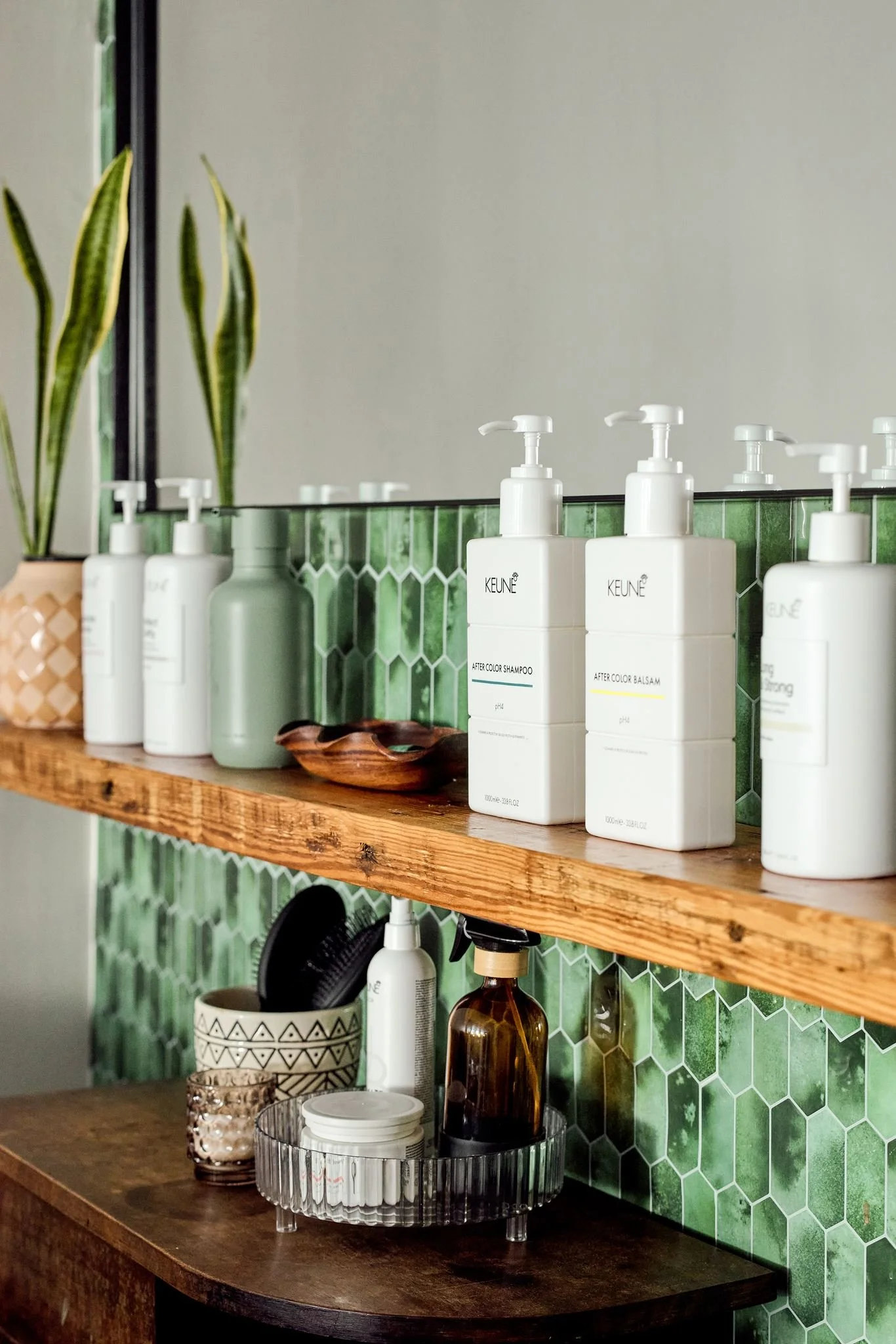Bathroom shelf with white bottles labeled 'Keune' for shampoo and balsam, a green bottle, a wooden dish, a decorative jar, and smaller containers, with a mirror and a plant with long green leaves reflected in the background.