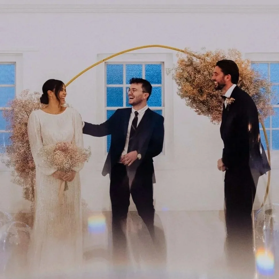 A wedding ceremony with a bride, groom, and officiant standing in front of a decorated backdrop; floral arrangements and a gold arch frame the scene.