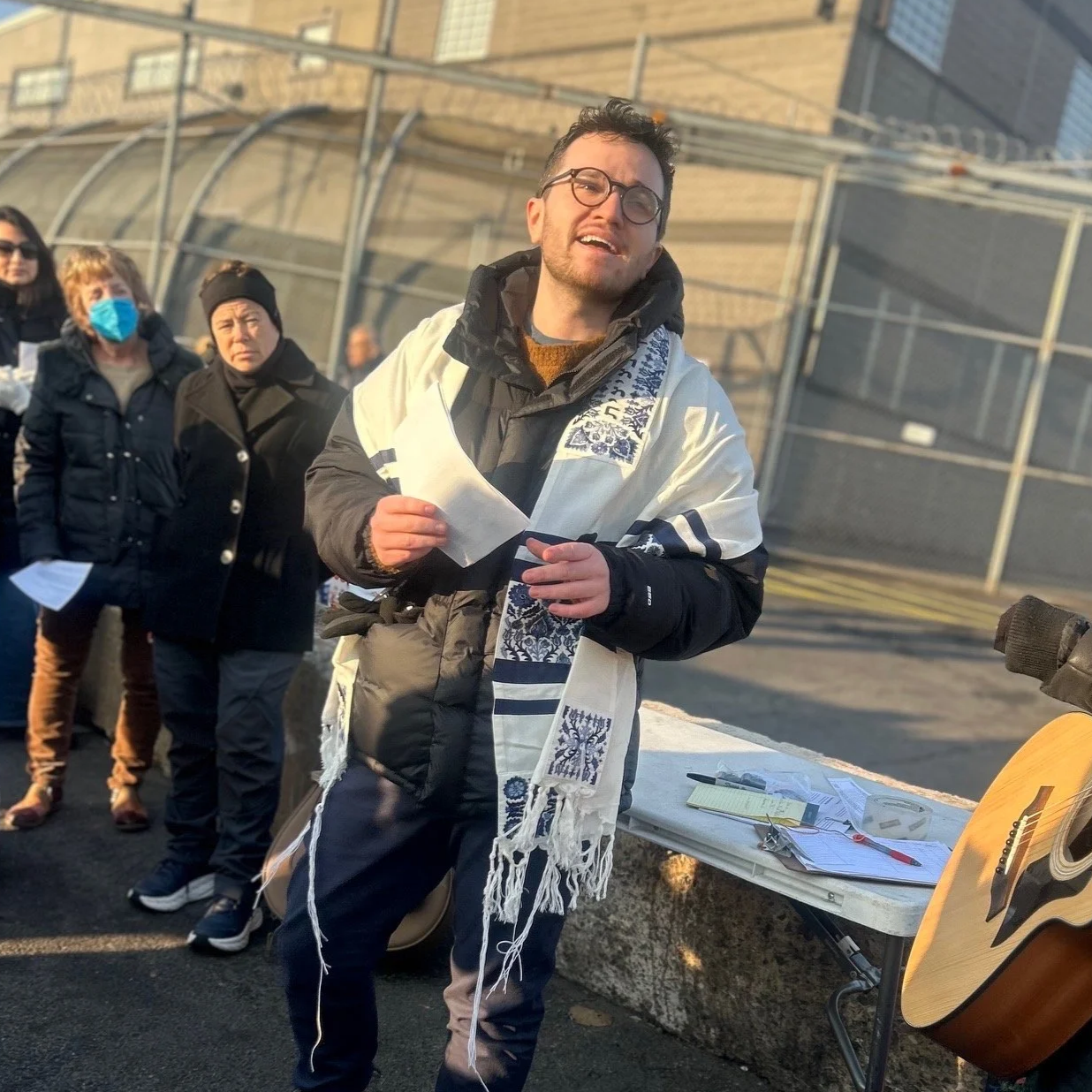 A young man with glasses and a beard standing in front of a group, speaking or singing, holding a piece of paper. He is wearing a black jacket, a black hoodie, and a white scarf with blue embroidery. There is a table with papers, a pen, and a guitar next to him, and several people standing behind him, some wearing masks.