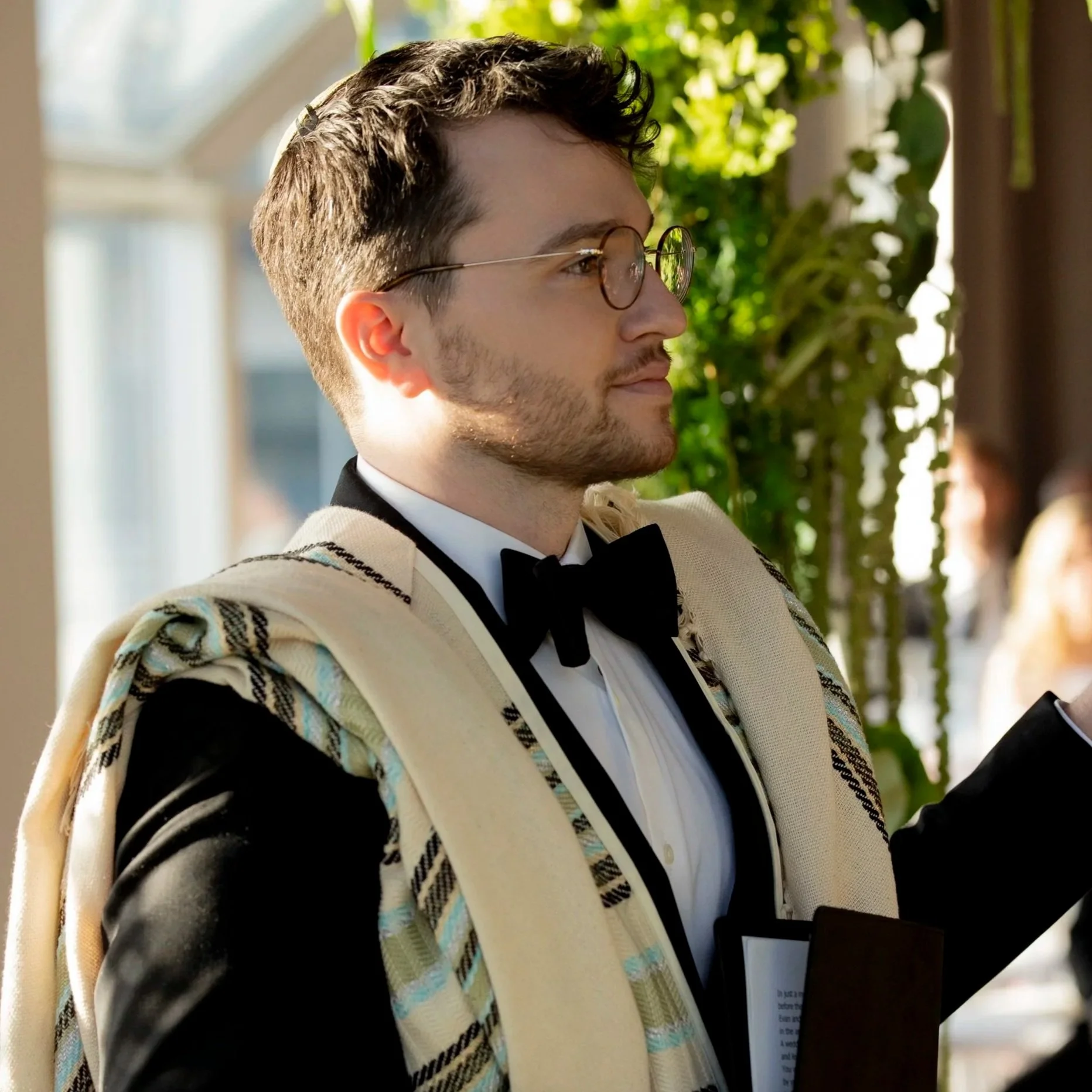 A man wearing round glasses and a tuxedo with a bow tie, sitting in a setting with sunlight and greenery in the background.