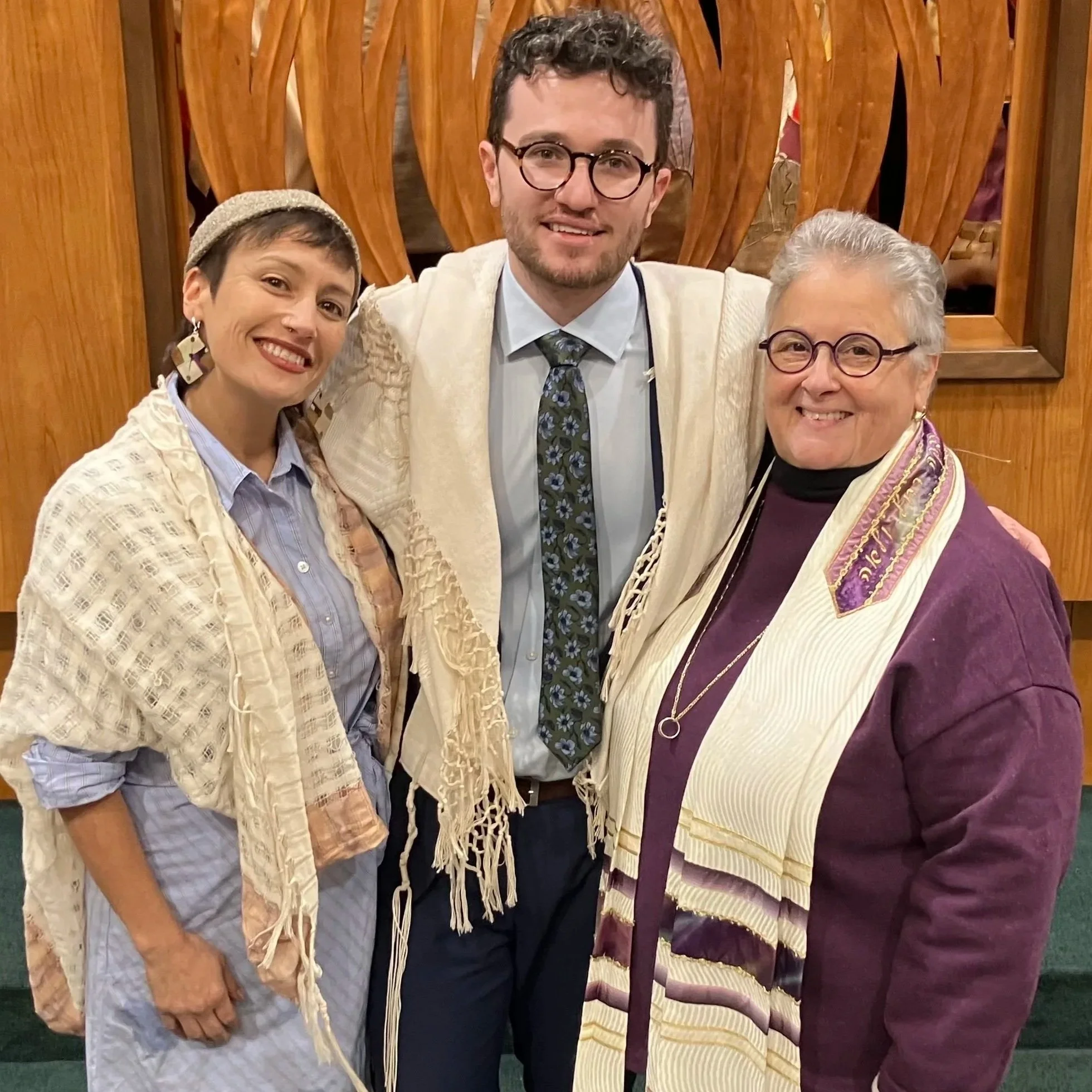 Three people standing close together in front of a wooden decorative wall, smiling. One person on the left is wearing a beige patterned shawl and a grey shirt. The person in the middle is wearing glasses, a white shirt, a floral tie, and a cream-colored knit sweater. The person on the right is wearing glasses, a maroon dress, and a cream-colored and purple shawl, and has short grey hair.