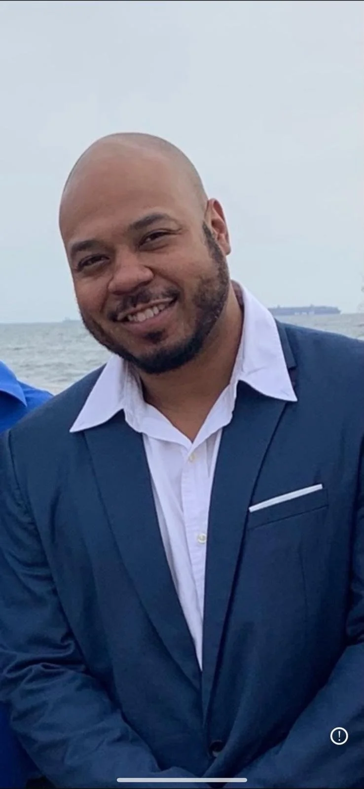 Smiling man in a dark suit and white shirt outdoors near water.