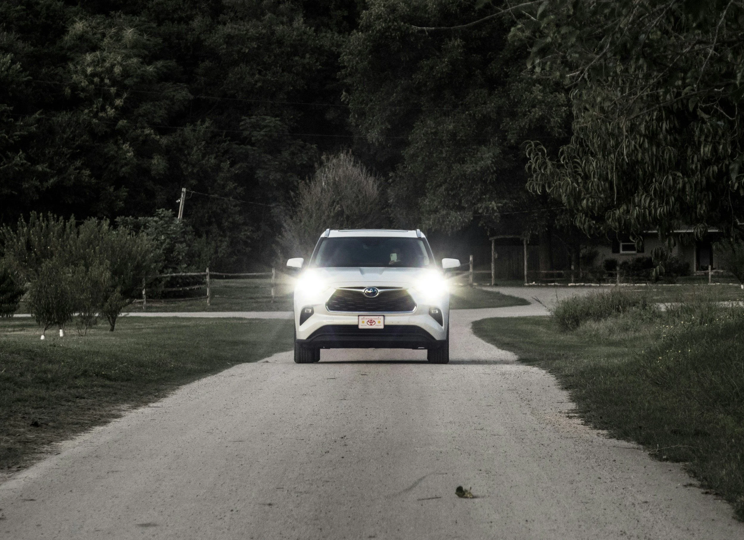 A white Toyota SUV parked on a gravel driveway at night, surrounded by trees and bushes with a dark sky overhead.