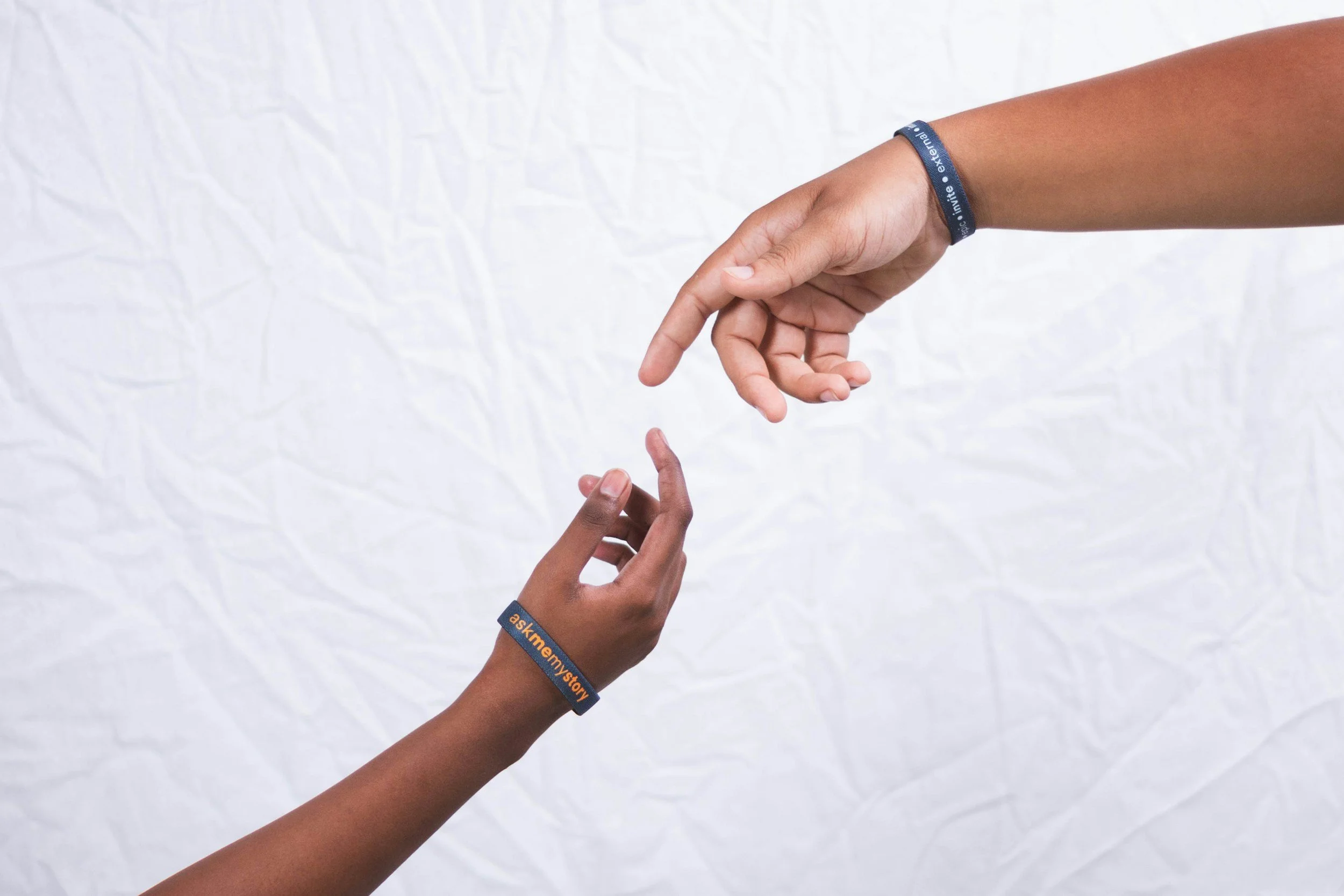 Two hands reaching towards each other with index fingers nearly touching, with a white crumpled background. Both wear black wristbands with orange and white text.