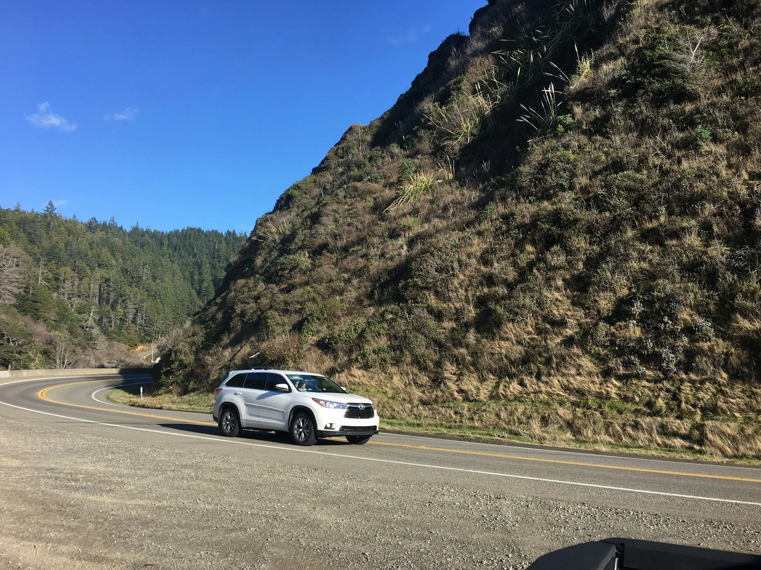 A white SUV parked on the side of a winding mountain road with a steep hillside covered in bushes and trees and a clear blue sky overhead.