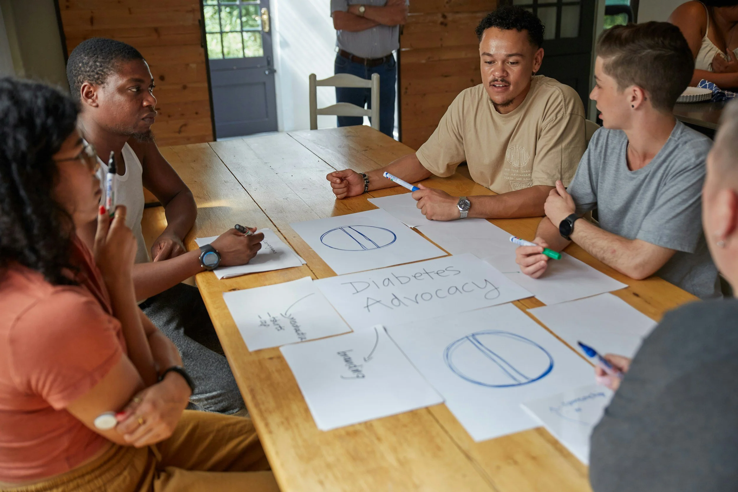 Group of people sitting around a wooden table during a meeting, with papers on the table including one with the words 'Diabetes Advocacy' and diagrams, in a room with natural light from windows.
