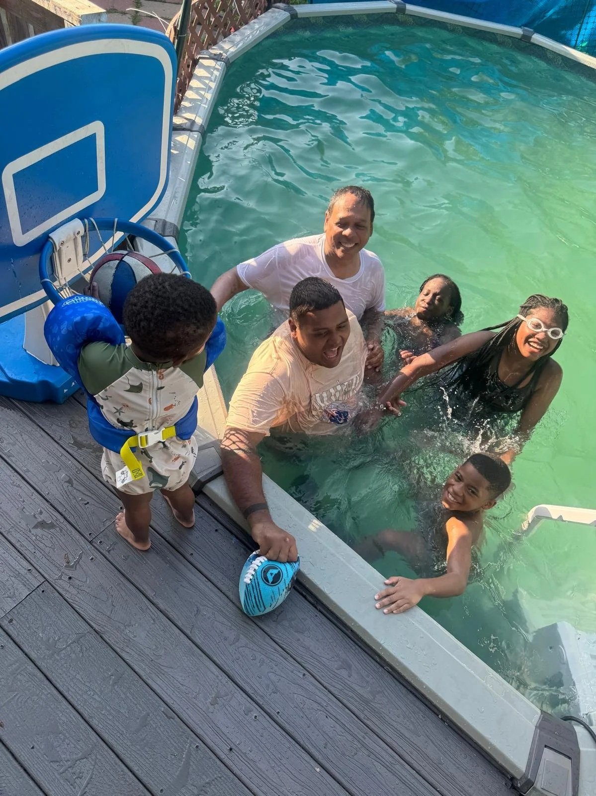 People enjoying swimming and smiling in a backyard pool, with a child standing on the pool deck.