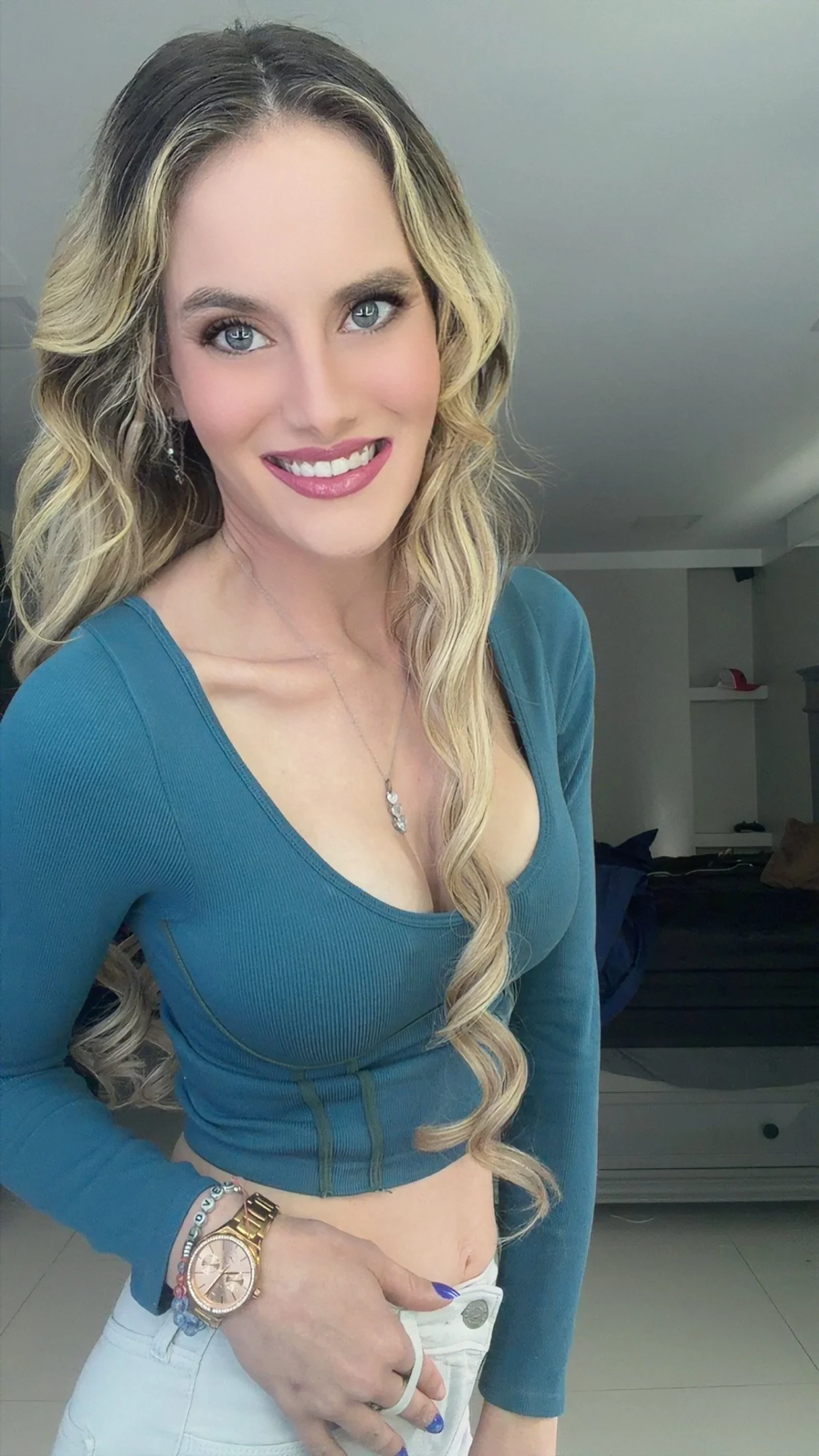 A woman with long blonde wavy hair, wearing a teal long-sleeve top, smiling and posing indoors, holding her waistband, with a watch and bracelets on her left wrist.