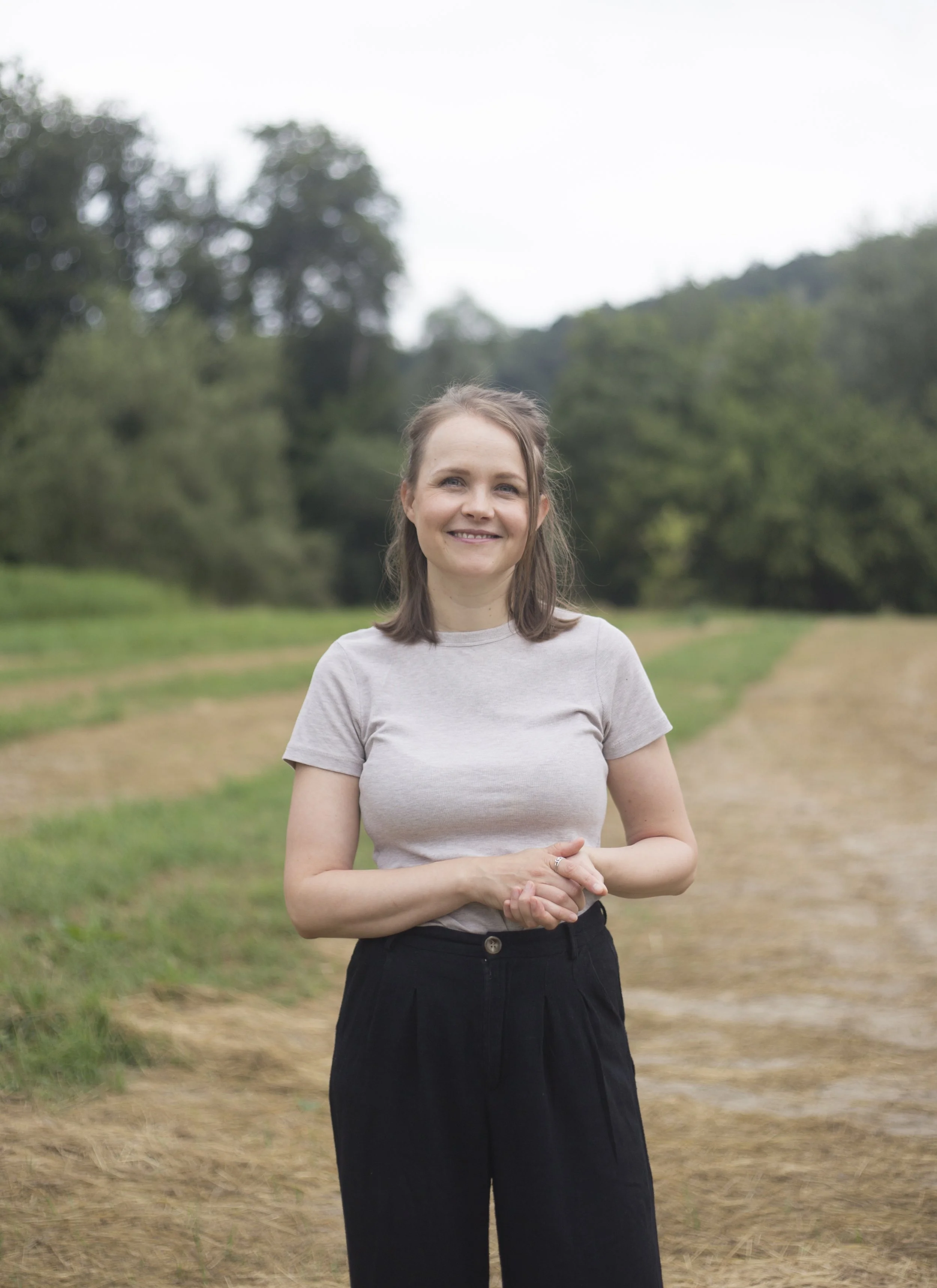 Eine lächelnde junge Frau steht auf einem Feld mit Bäumen im Hintergrund, trägt ein graues T-Shirt und schwarze Hosen.