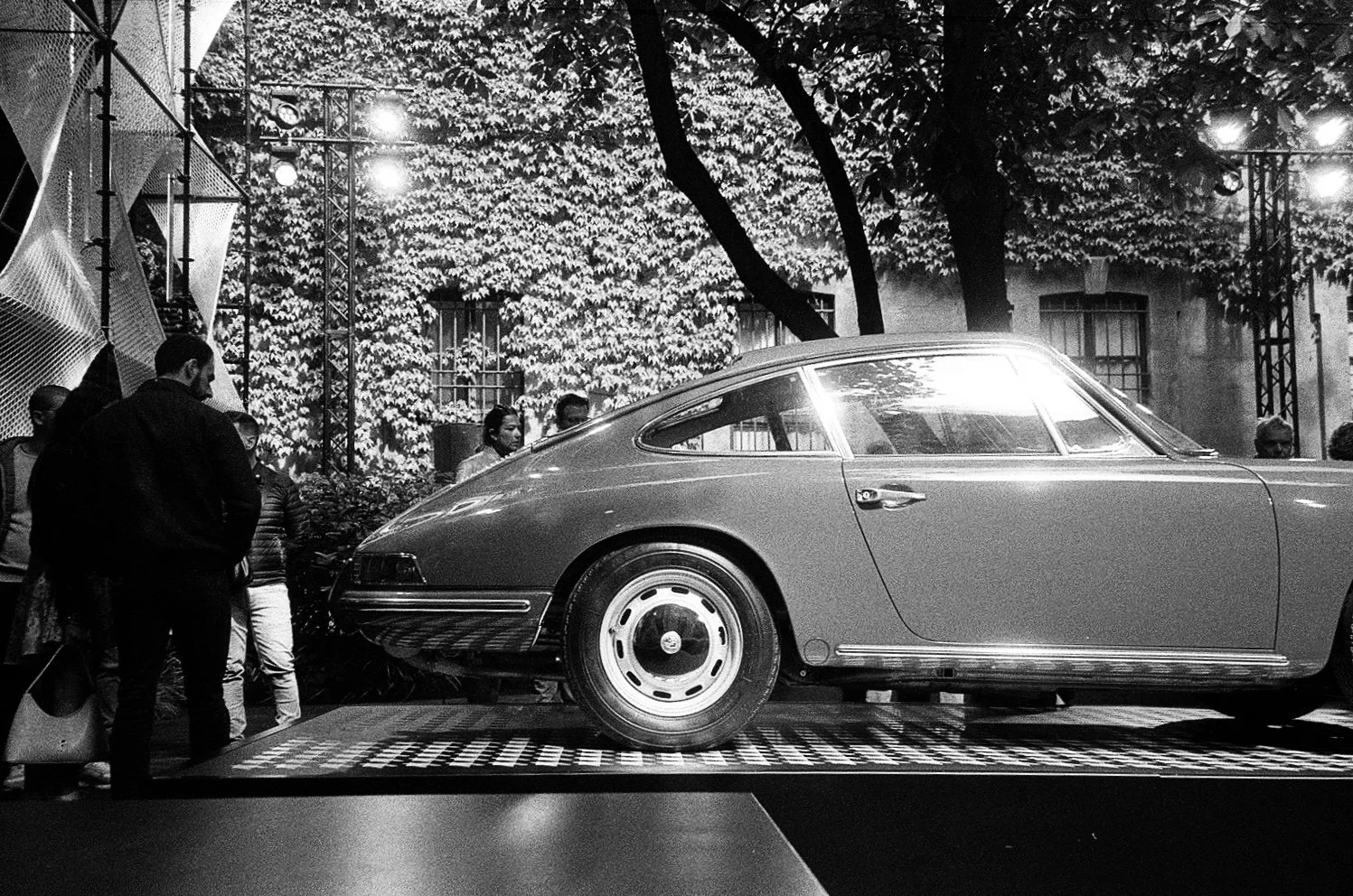 Auto d'epoca esposta su pedana in un evento di auto, con persone intorno che la osservano, sullo sfondo un edificio ricoperto di foglie e luci nel pomeriggio o sera.