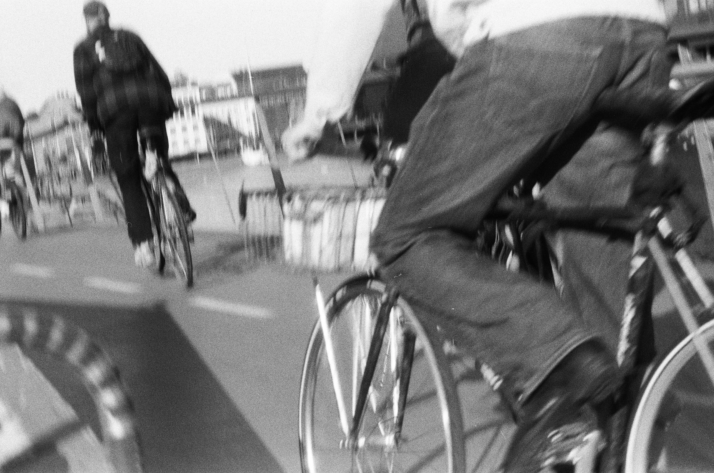 Foto in bianco e nero di persone in bicicletta su una strada urbana, con edifici sullo sfondo
