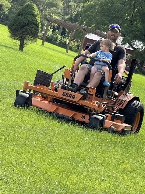 A man and a young boy riding a large orange zero-turn lawn mower on a grassy lawn with trees and a small red building in the background.