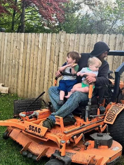 A person with a hood sitting on a large orange lawn mower with two young children, one sitting on their lap and the other sitting beside them, in a backyard with a wooden fence and trees in the background.