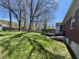 Backyard with a lush green lawn, leafless trees, a wooden deck with stairs, and a brick house with a window, under a clear blue sky.