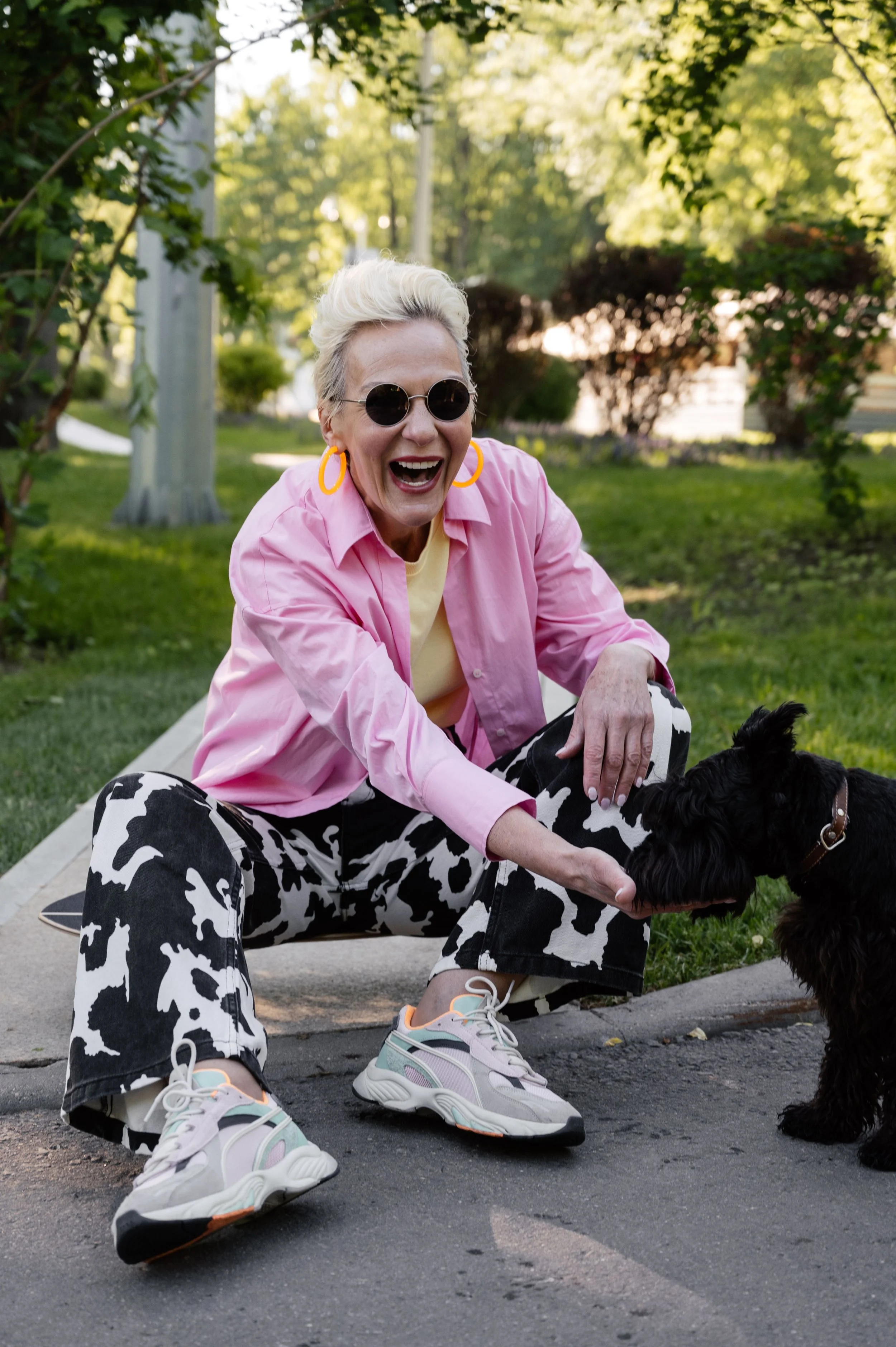 Woman wearing sunglasses, a pink shirt, cow-print pants, and sneakers sitting outdoors with a black dog.