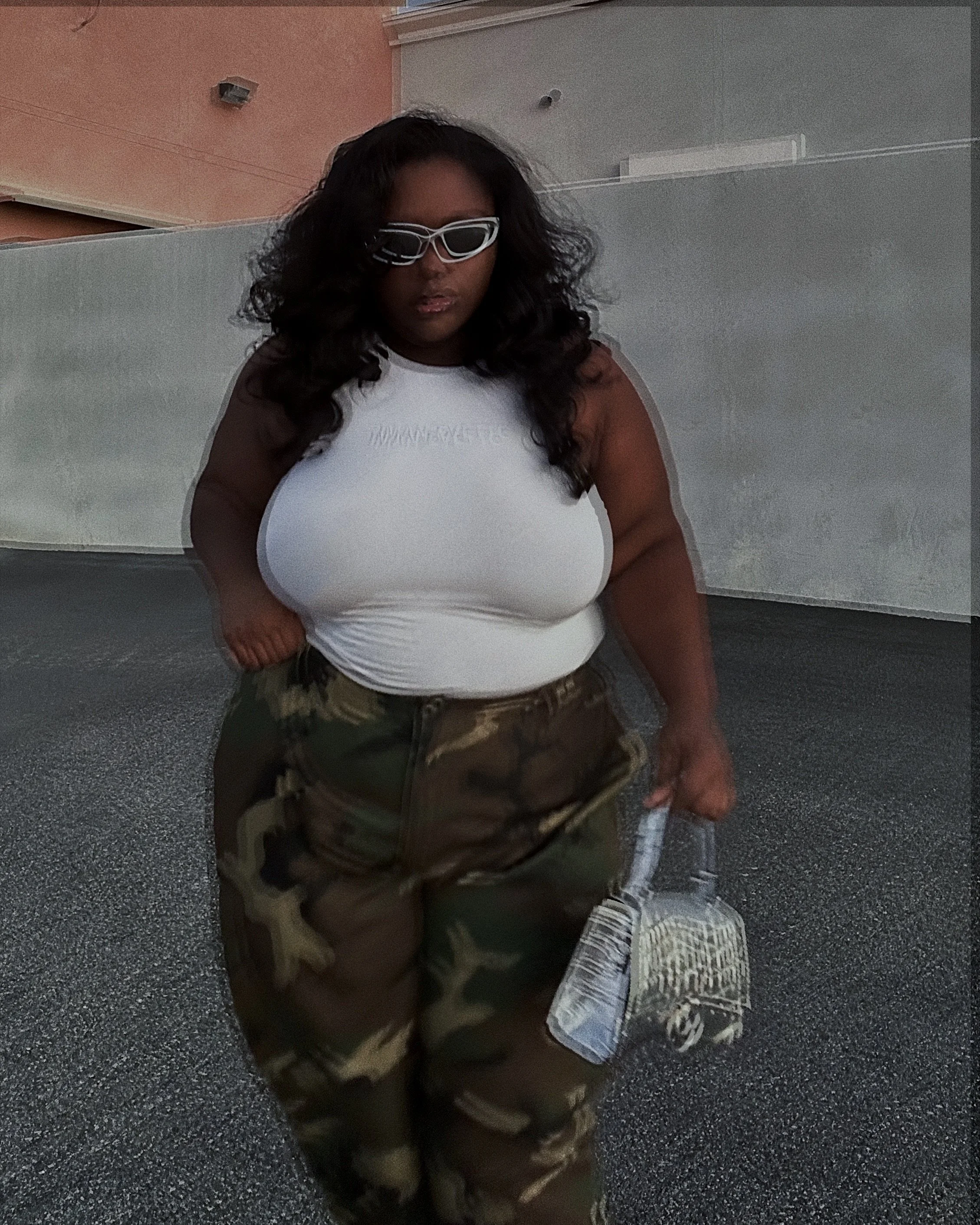Woman wearing white sunglasses, a white sleeveless top, and camouflage pants, seated indoors.