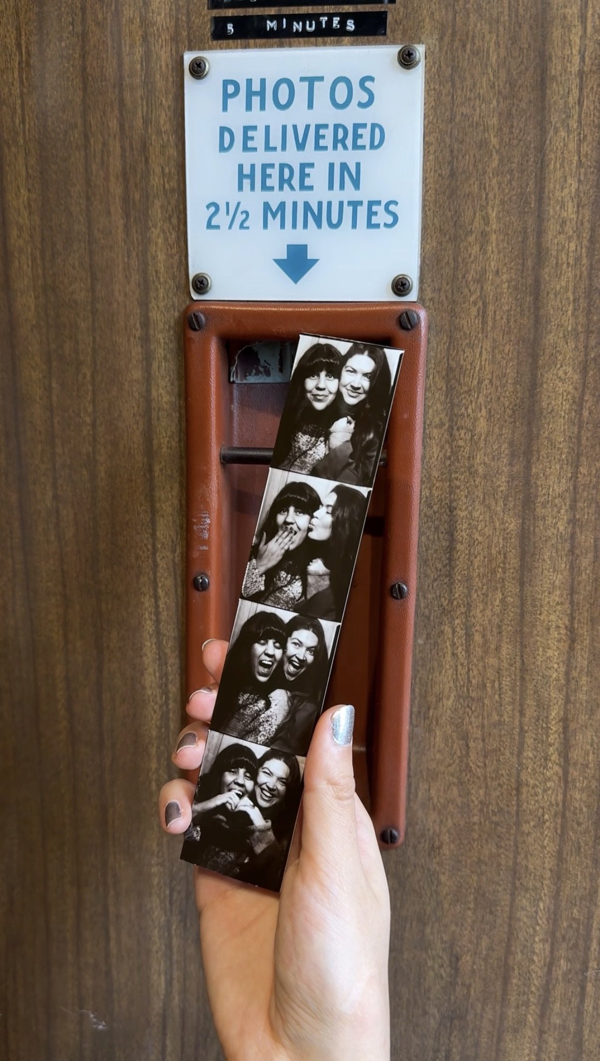 Hand holding a black-and-white photo booth strip beneath a sign promising photos delivered in 2½ minutes.