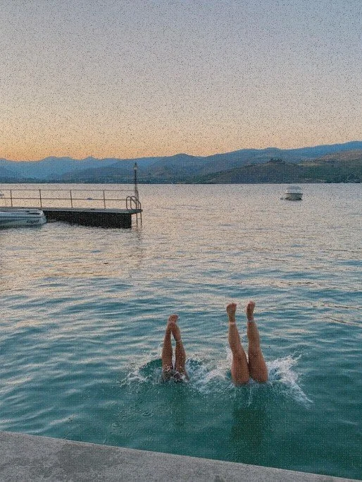 Two swimmers diving feet-first into a lake at sunset, with a dock, boat, and mountains in the background