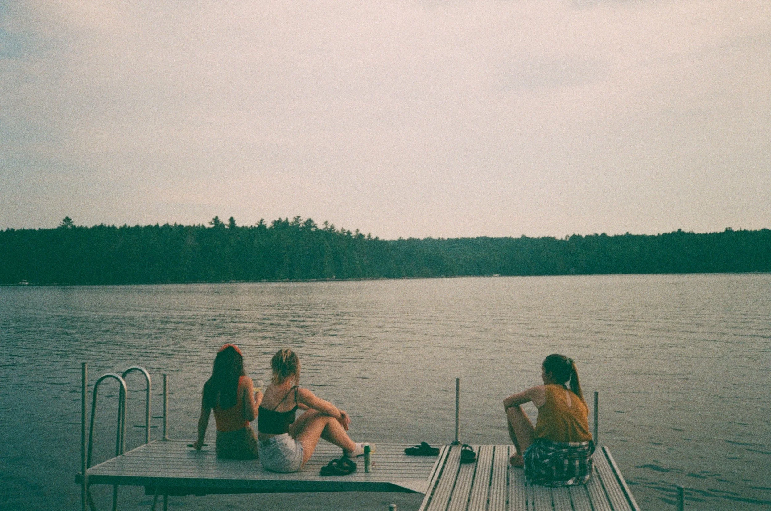 Three women relaxing on a dock over a lake with a forested shoreline