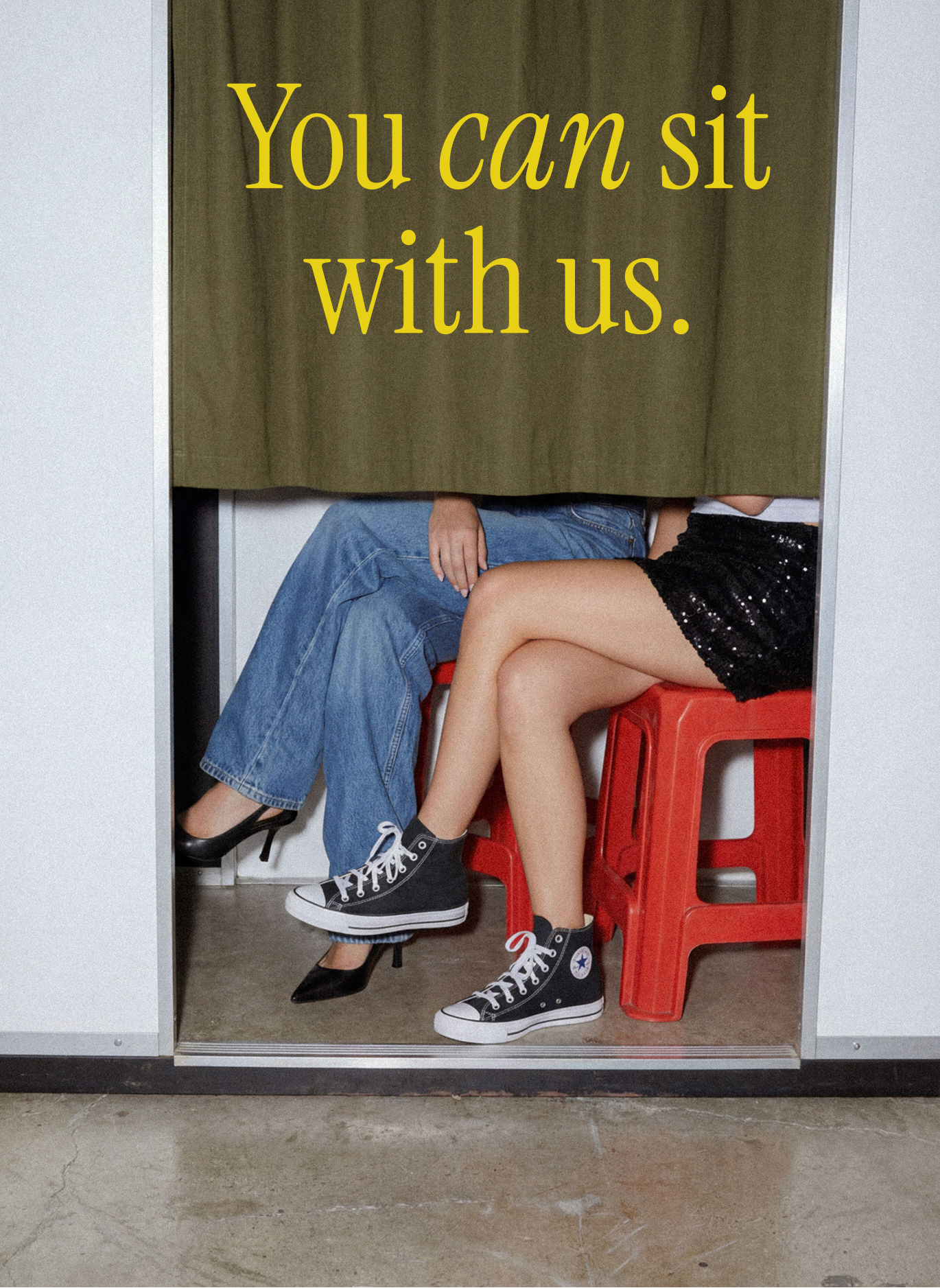 Two people sitting behind a green curtain on red stools, wearing jeans, black heels, and black Converse sneakers, with yellow text reading “You can sit with us.”