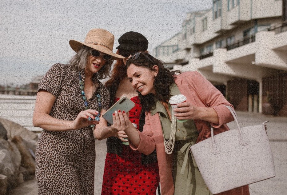 Three stylish women in sun hats look at a smartphone outdoors near a waterfront building.