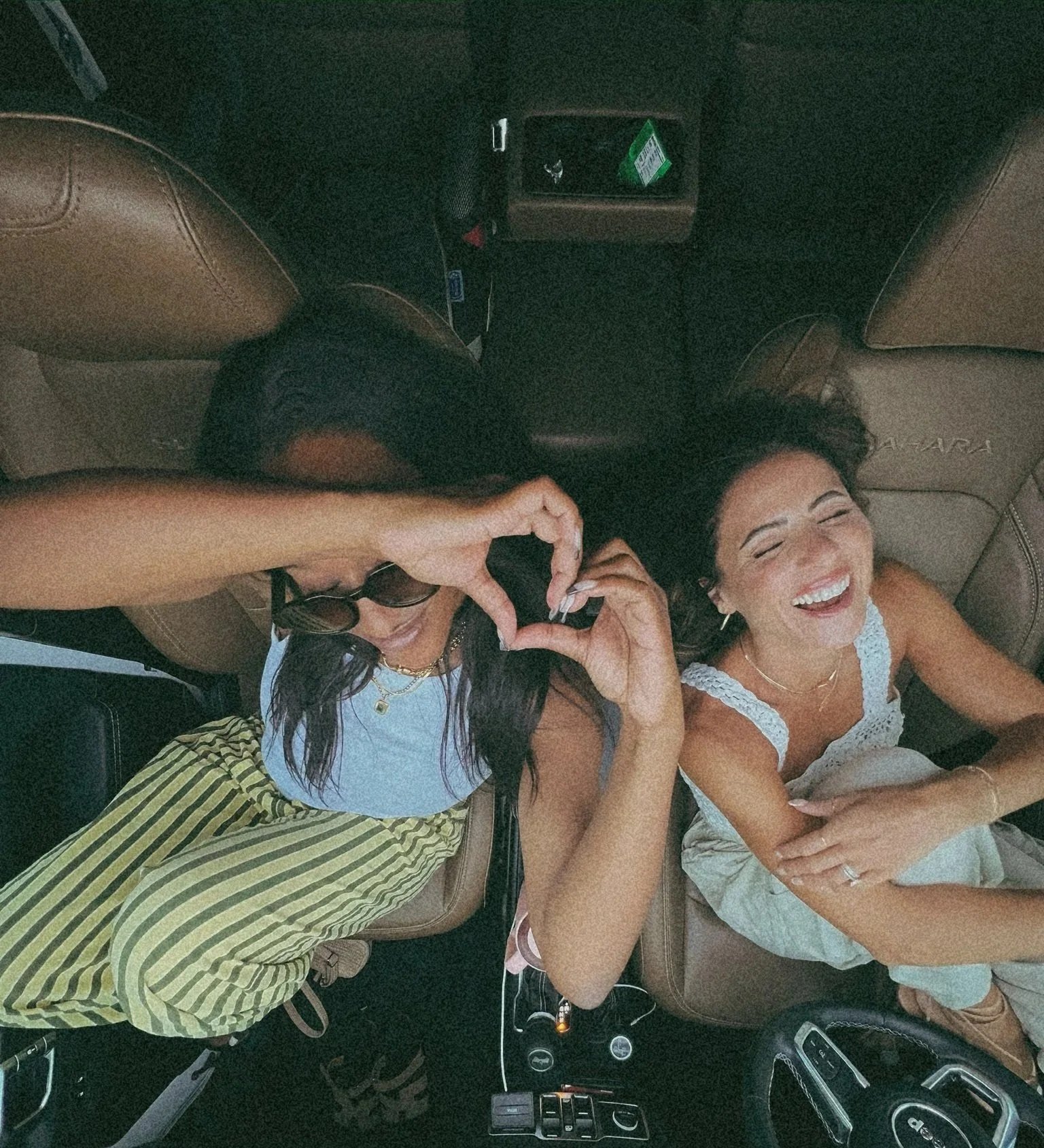 Two women laughing and making a heart shape with their hands inside a car.