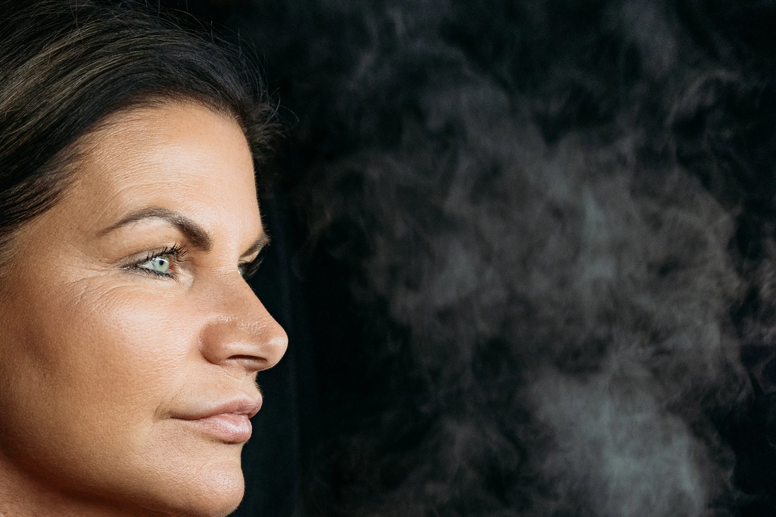 Close-up of a woman's face with dark hair, showing her eye, eyebrow, and the side of her nose against a black background.