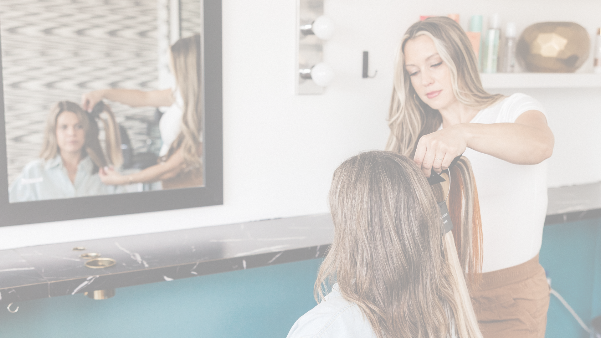 A woman with long, wavy hair and a white shirt is giving a haircut or styling a person's hair. The background shows shelves with colorful hair products and a mirror with lights.