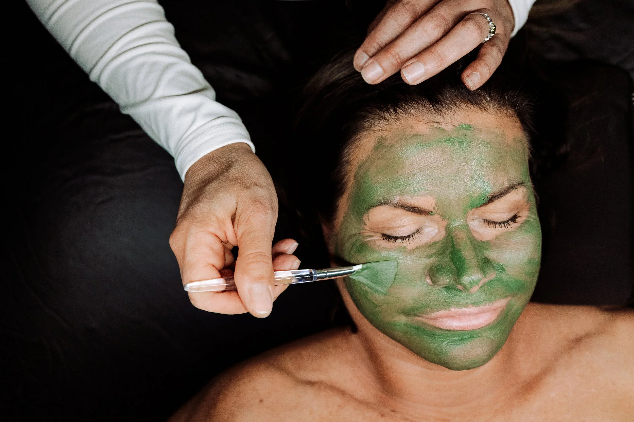 Person receiving a green facial mask while lying down with eyes closed, dark hair, and hands gently touching their head.