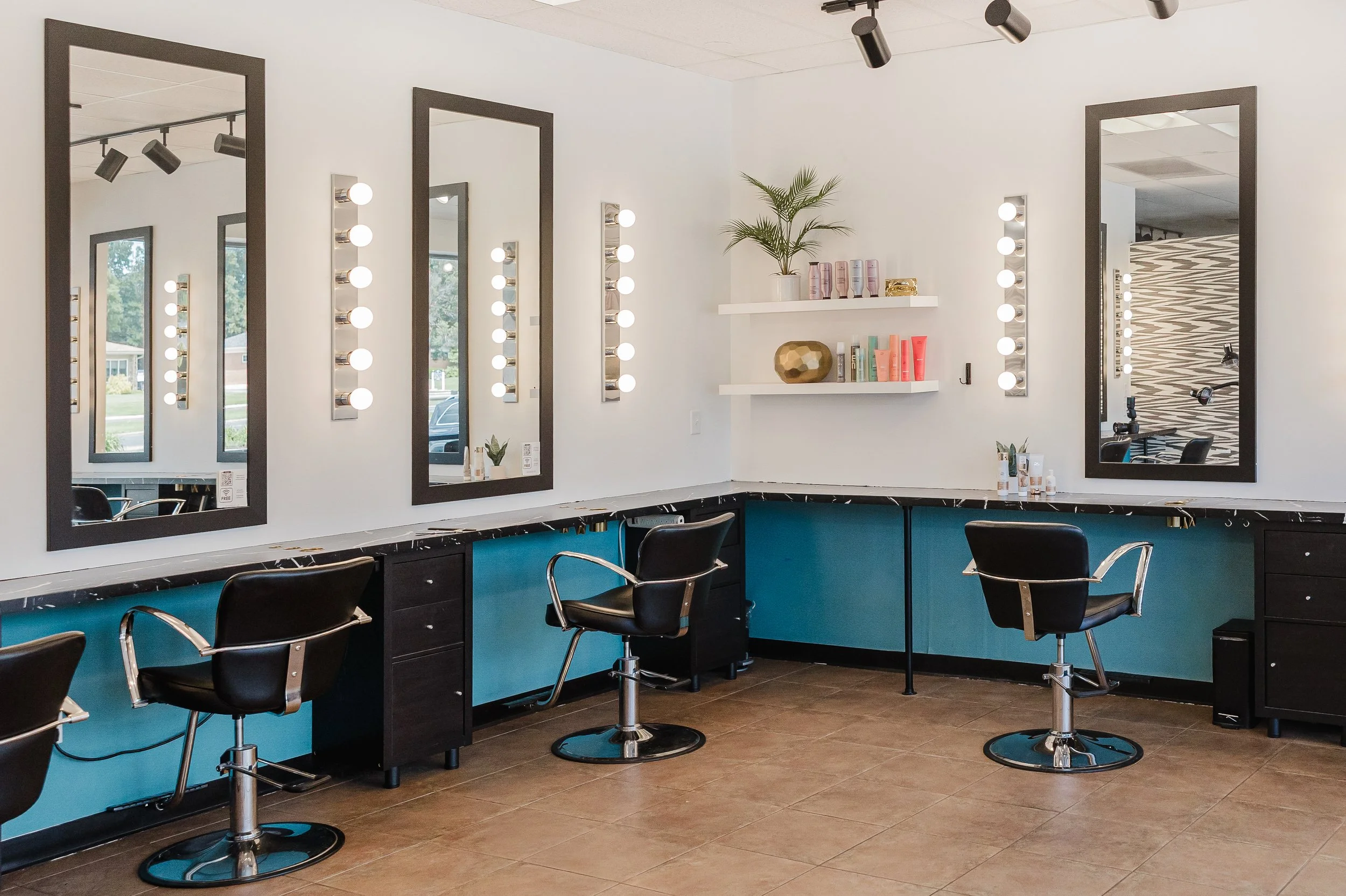 A beauty salon with three large mirrors with black frames, round vanity lights, black chairs, a blue counter, and shelves with hair care products. There's a plant and decorative items on the shelves.