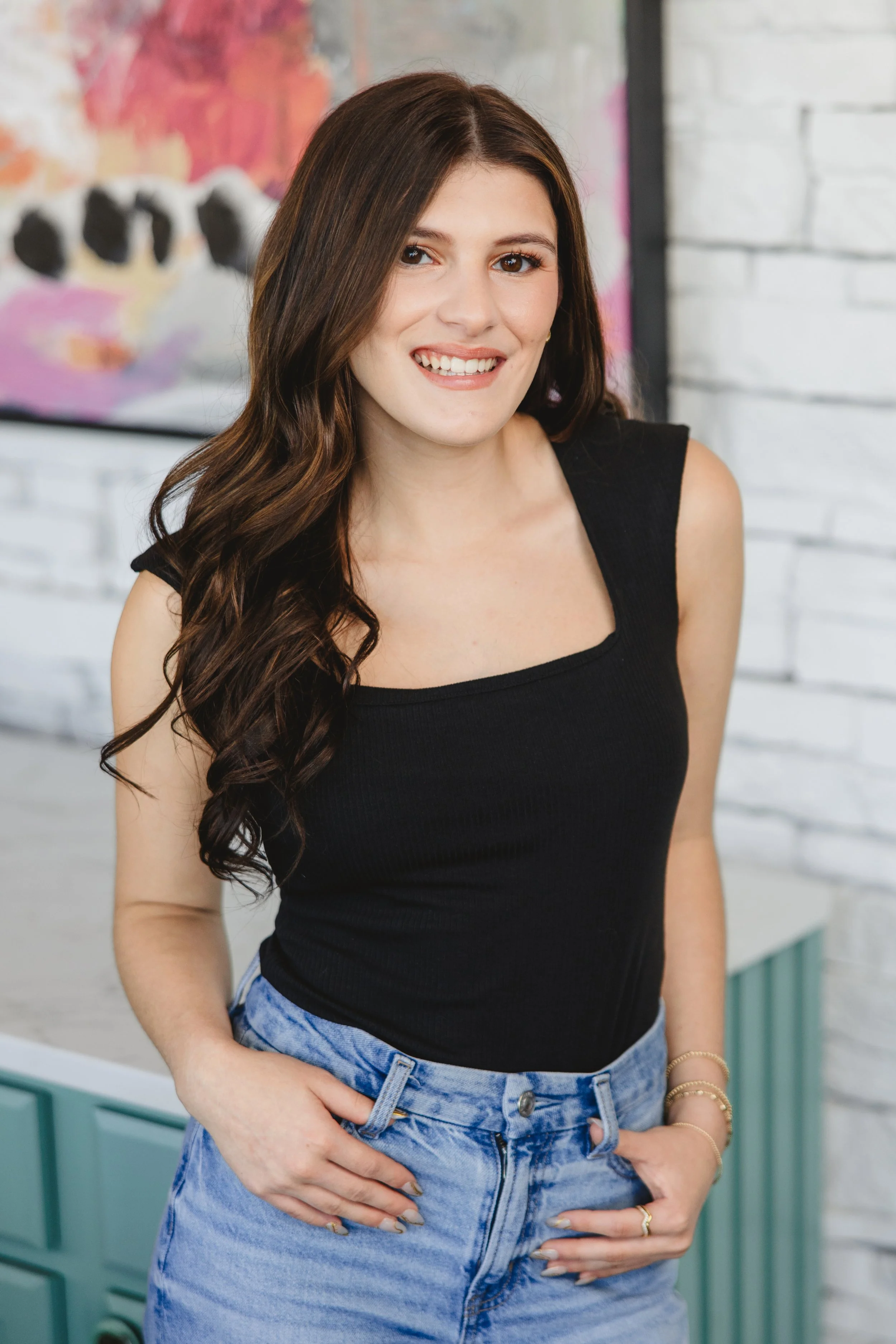 A woman with long, wavy brown hair smiling, wearing a black sleeveless top and high-waisted light blue jeans, with a colorful abstract painting and white brick wall in the background.