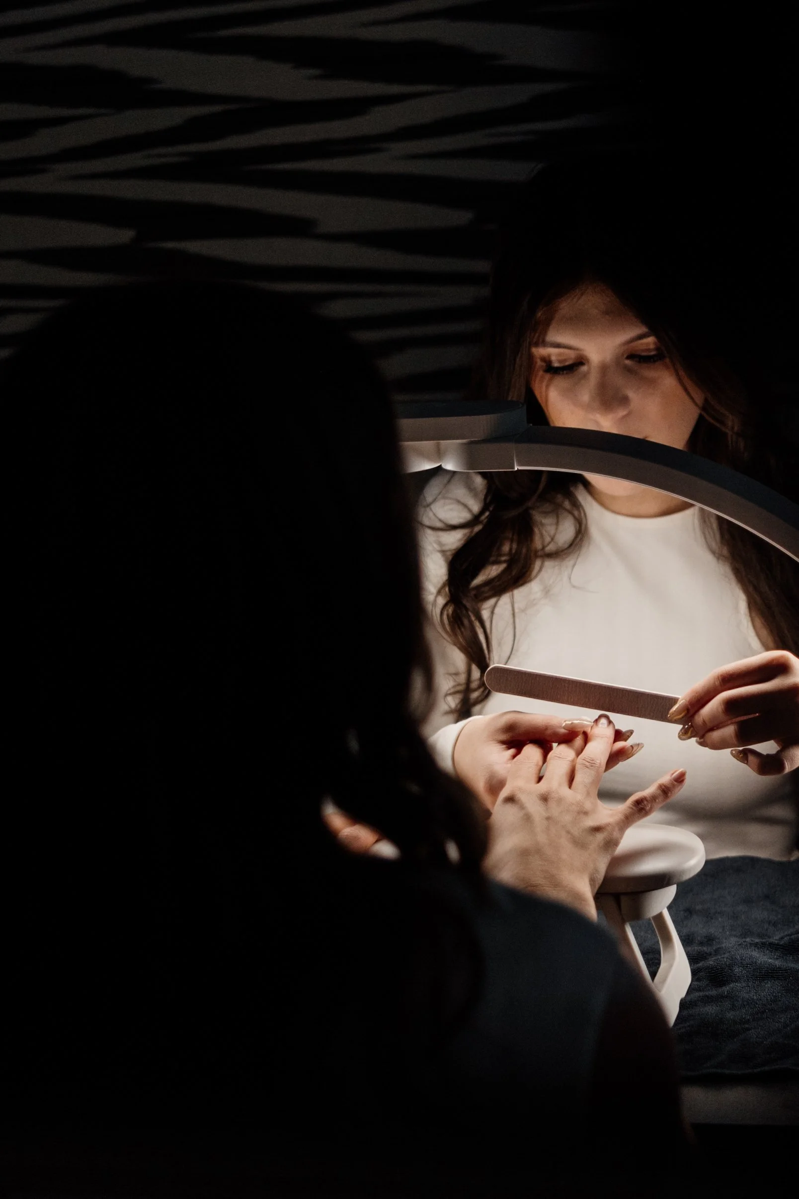 A woman with wavy brown hair looking down at her nails, which are being painted by someone else, under a curved overhead light in a dark environment with horizontal striped ceiling.