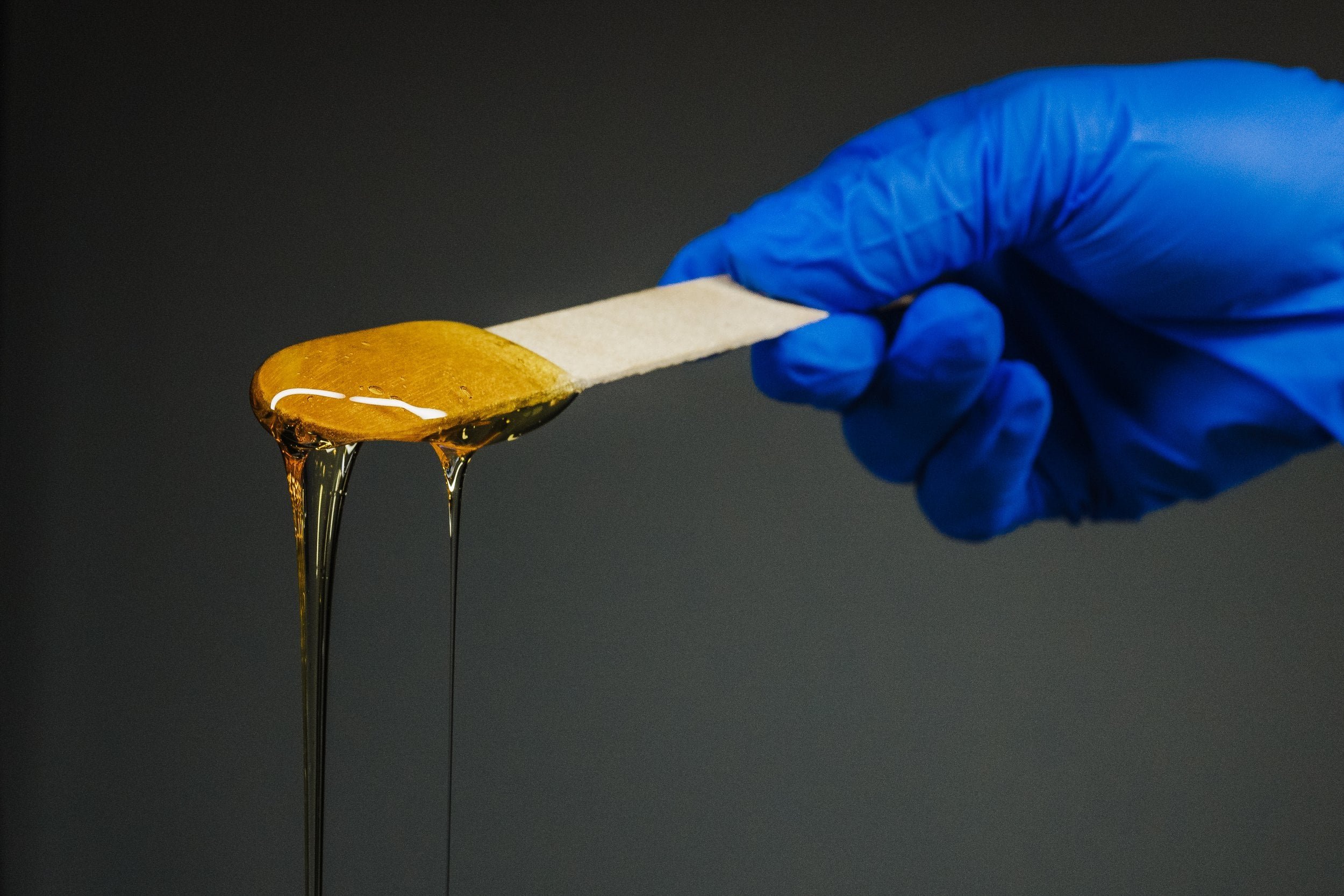 A hand wearing a blue glove holding a wooden spatula with honey dripping from it.