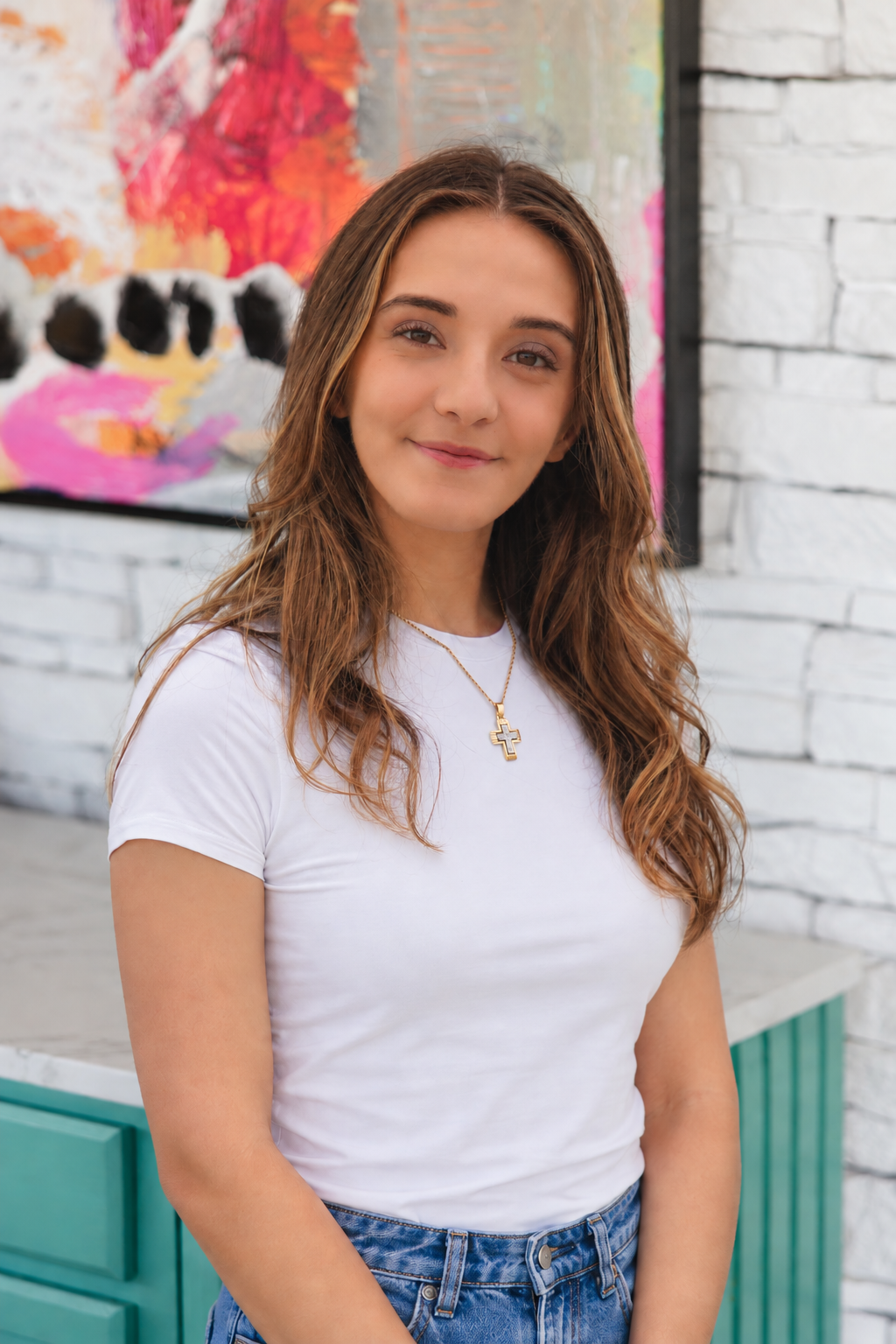 Portrait of a young woman with long, wavy brown hair, wearing a white t-shirt and a gold cross necklace, standing in front of an abstract colorful painting on a white brick wall.
