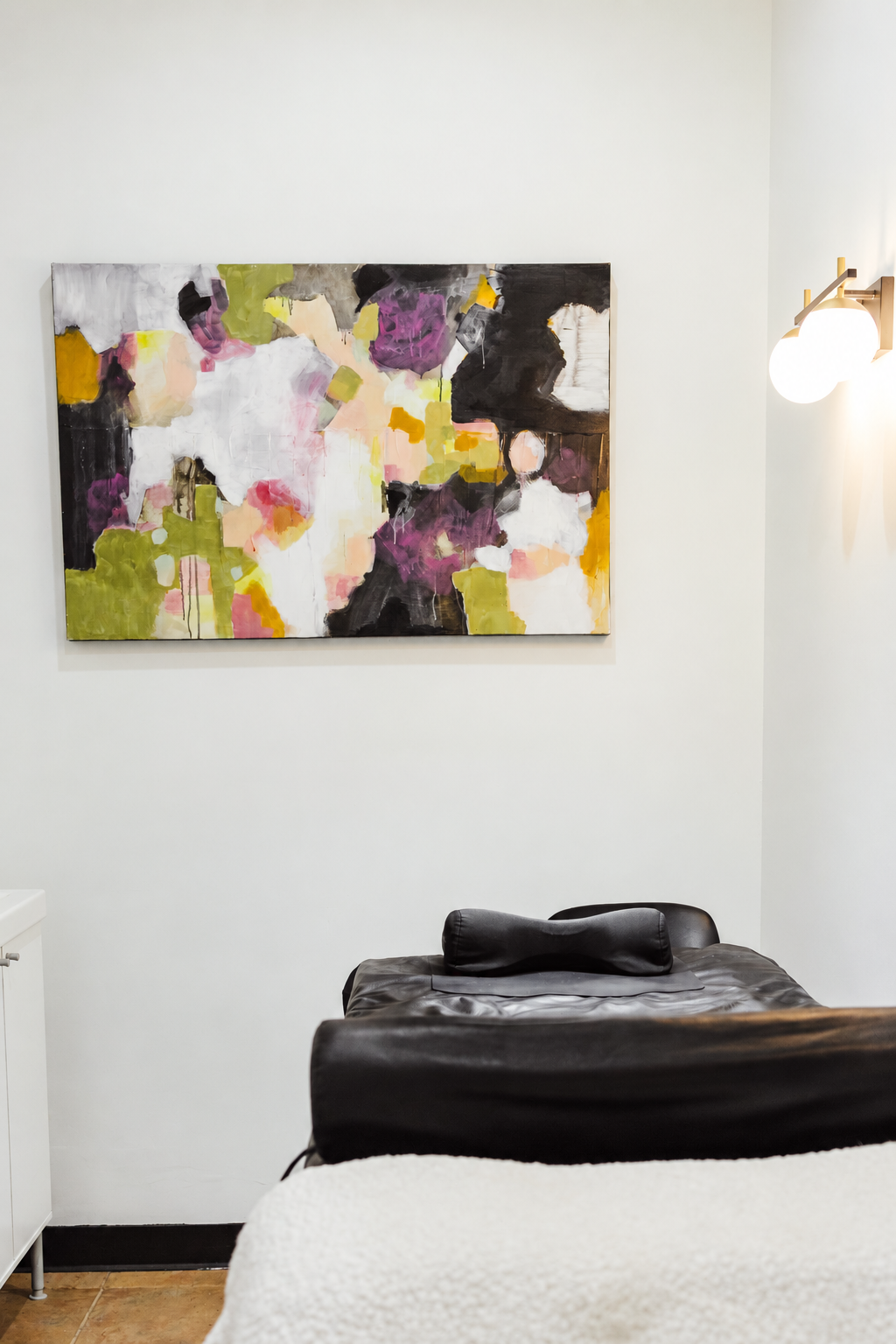 Abstract colorful painting on a white wall in a room with a black massage bed, a white side table, and a wall sconce with two round bulbs.