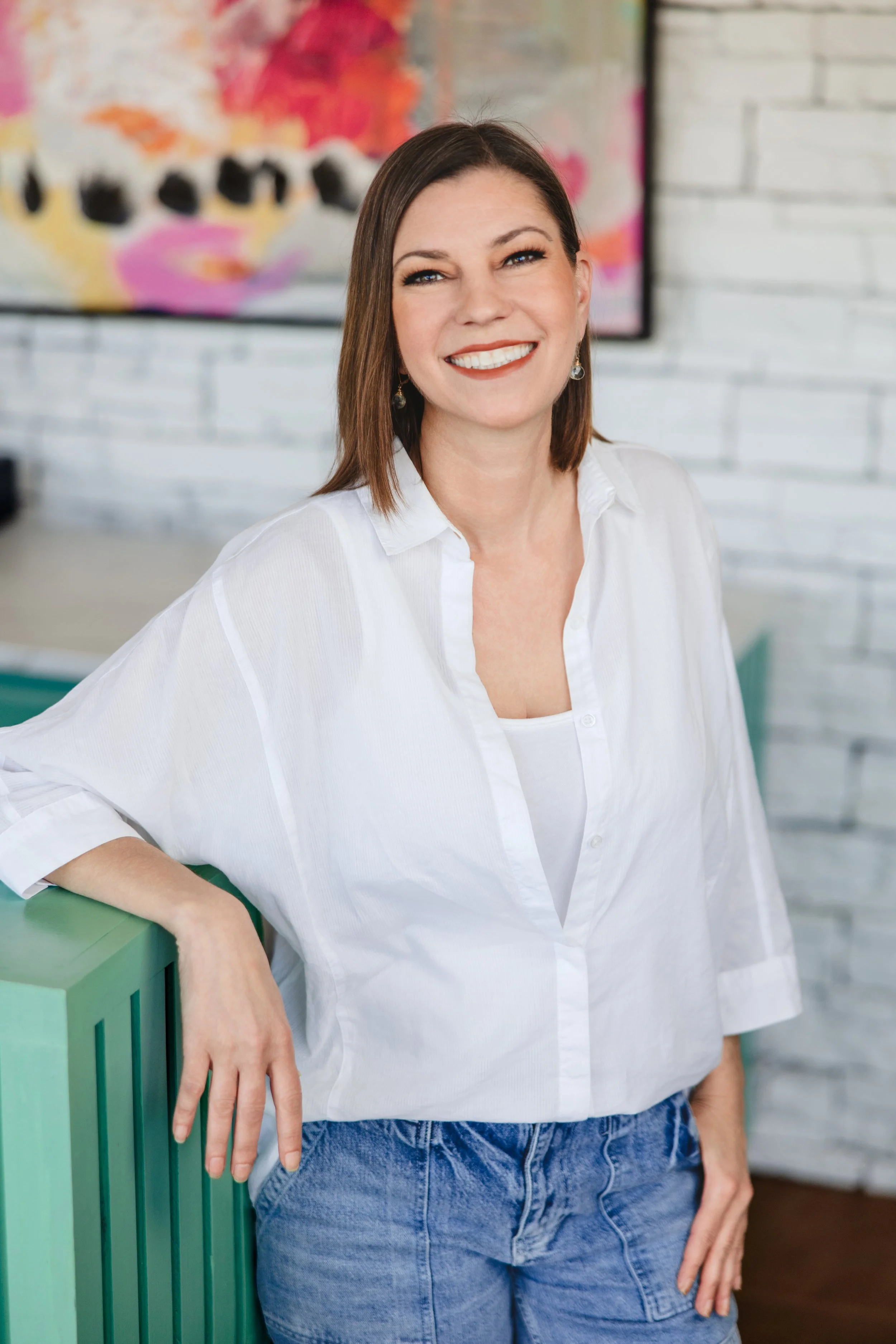 A woman wearing a white shirt and blue jeans, standing and leaning on a green railing in an indoor setting with a colorful blurred abstract painting and white brick wall in the background, smiling at the camera.