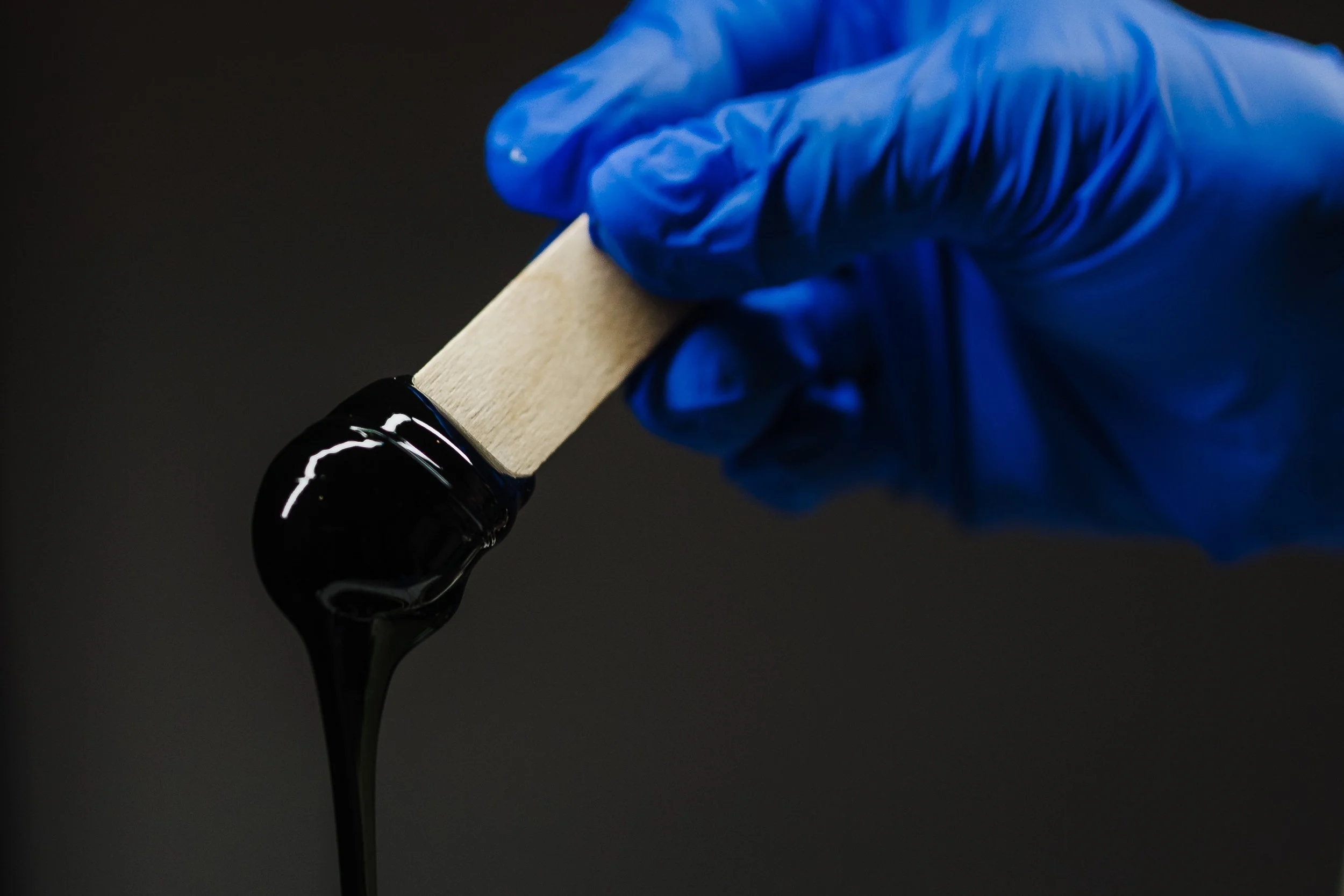 A gloved hand holding a wooden stick pouring wax against a black background.