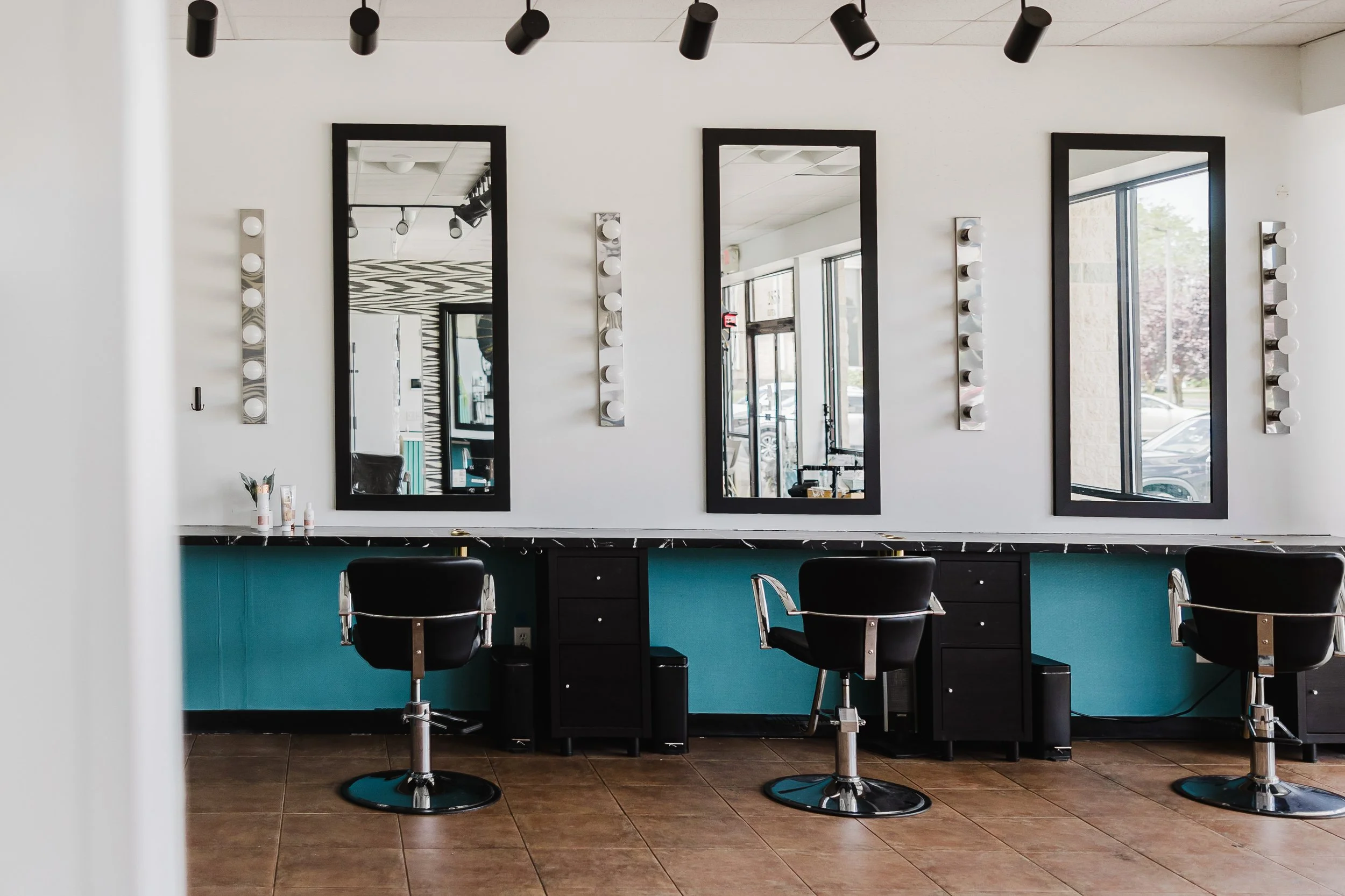 View of a modern hairstyling station with three mirrors framed in black, styled with white bulbs on either side, black chairs with chrome bases, a turquoise counter below, and items like bottles on a white shelf. The room has large windows and black ceiling spotlights.
