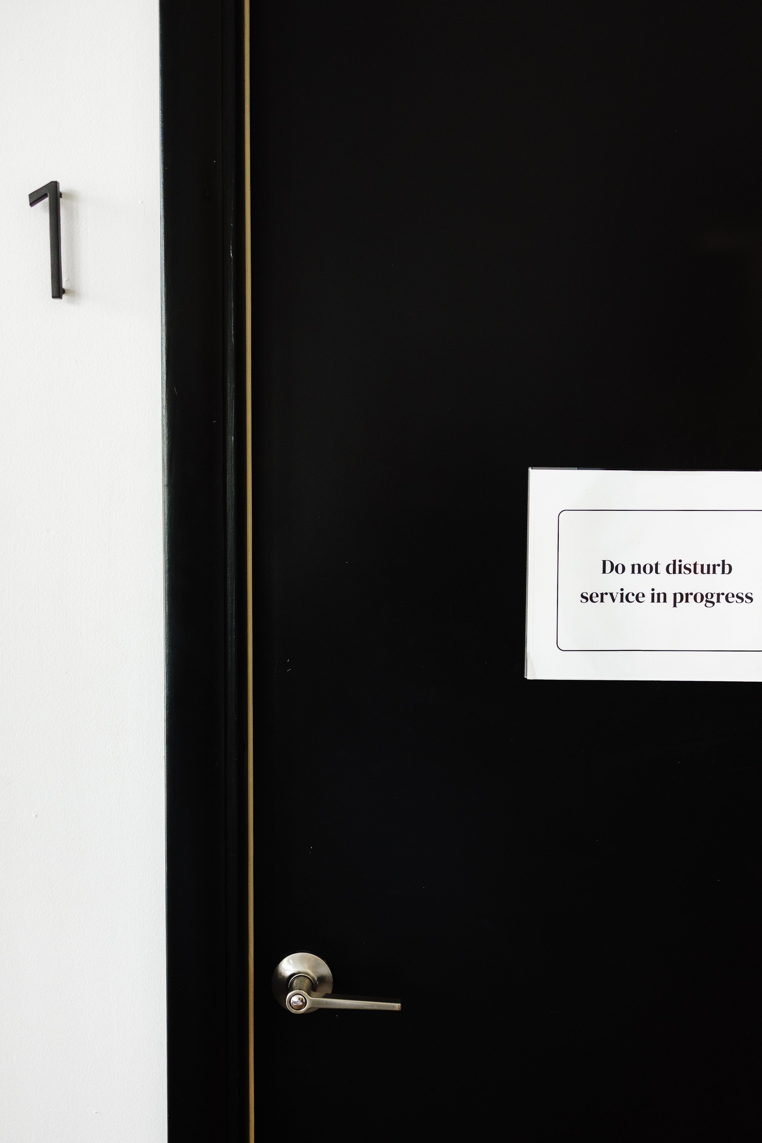 Black door with a silver handle and a white sign that reads 'Do not disturb service in progress'. There is a number 1 on the wall next to the door.