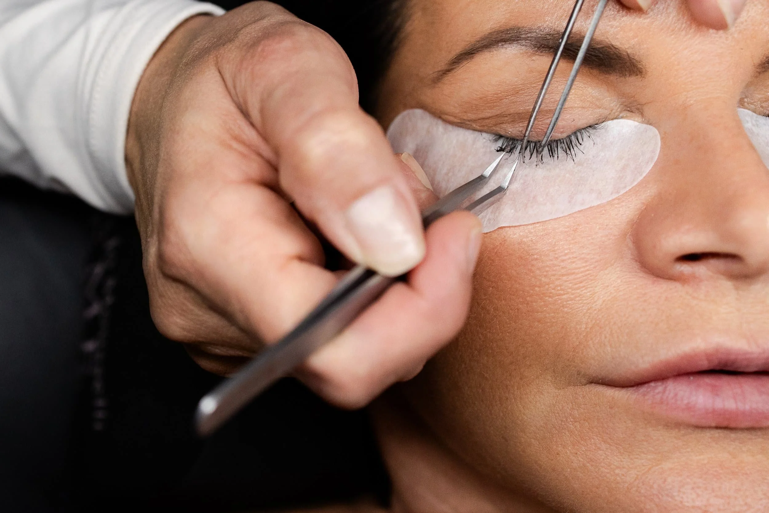 Close-up of a person receiving an eyelash extension procedure, with a technician using tweezers to attach artificial lashes to the person's natural lashes.